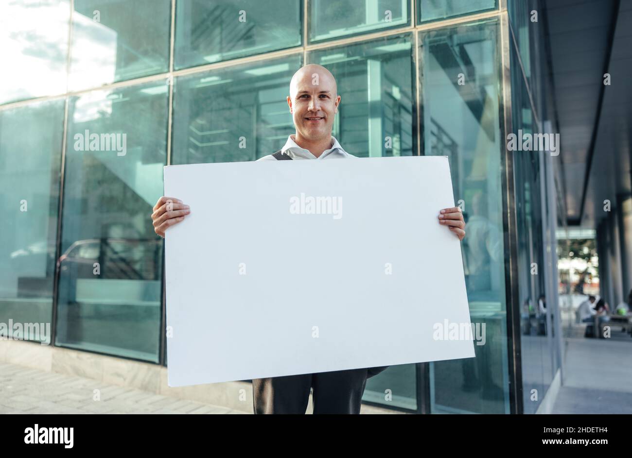 Fröhlicher Geschäftsmann mit einem leeren Banner und einem Lächeln in die Kamera, während er vor einem hohen Bürogebäude in der Stadt steht. Geschäftsakt Stockfoto