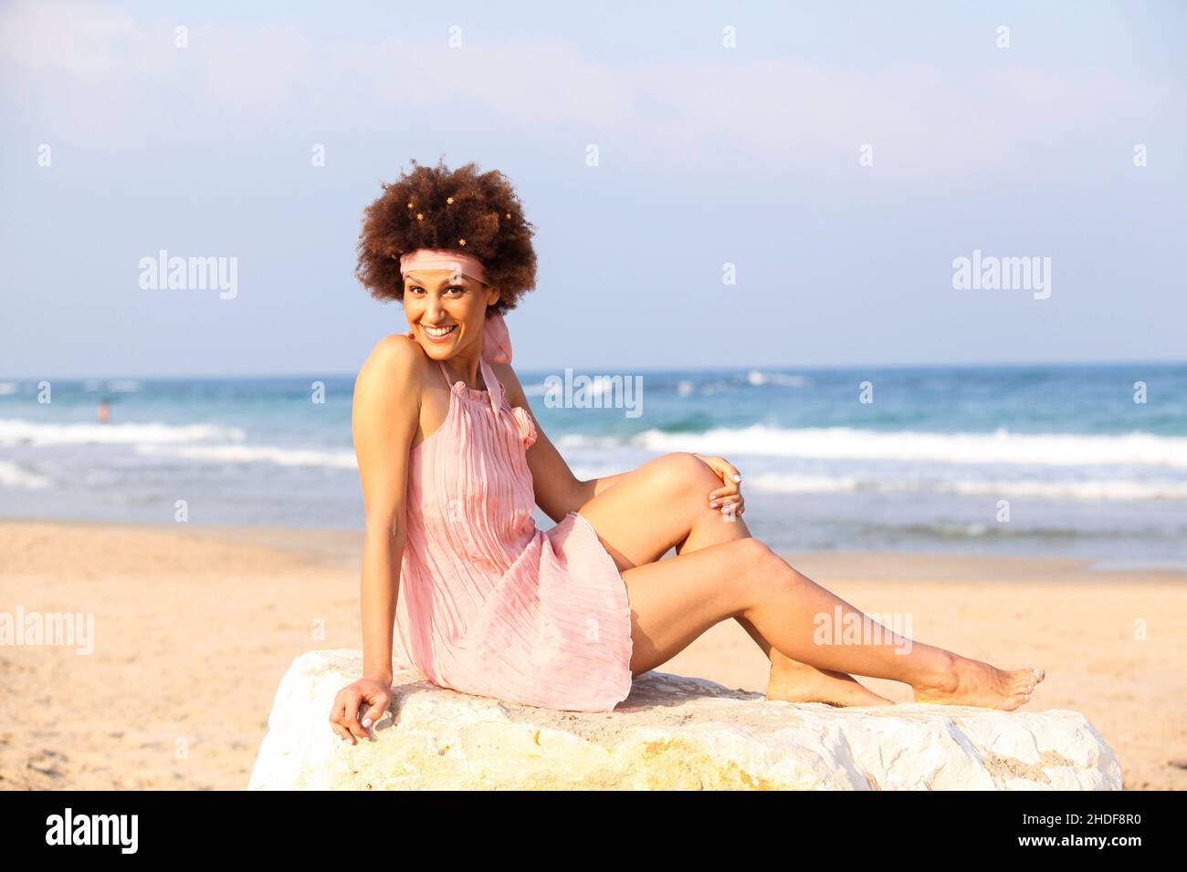 Eine fröhliche, lächelnde 28-jährige Frau im New Age-Stil sitzt auf einem Felsen am Strand Stockfoto