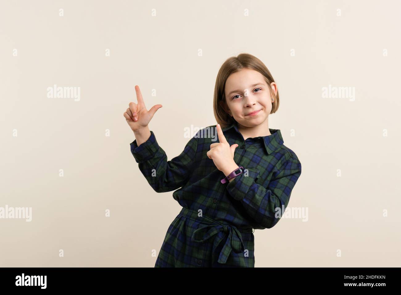 Cute Little Girl 9s in karierten Hemdkleid zeigt zwei Finger in beige Hintergrund. Kindsfinger, die auf den Index zeigen, kopieren den Platz für die Anzeige. Wow, schau, wirb hier Werbung. Stockfoto