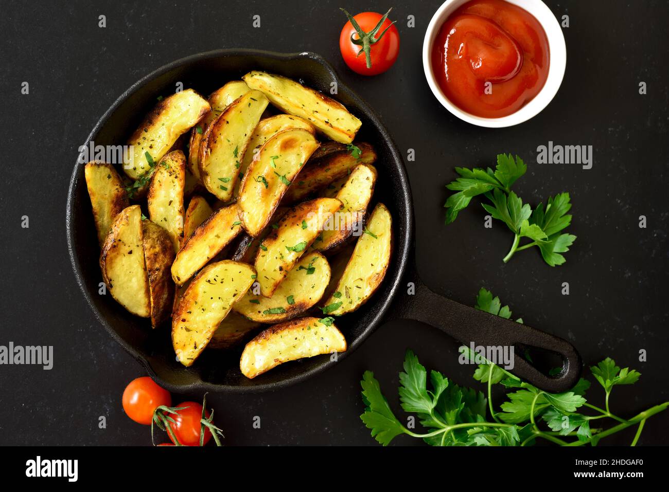 Gebratene Kartoffelkeile in einer Pfanne auf schwarzem Steingrund. Draufsicht, flach liegend Stockfoto