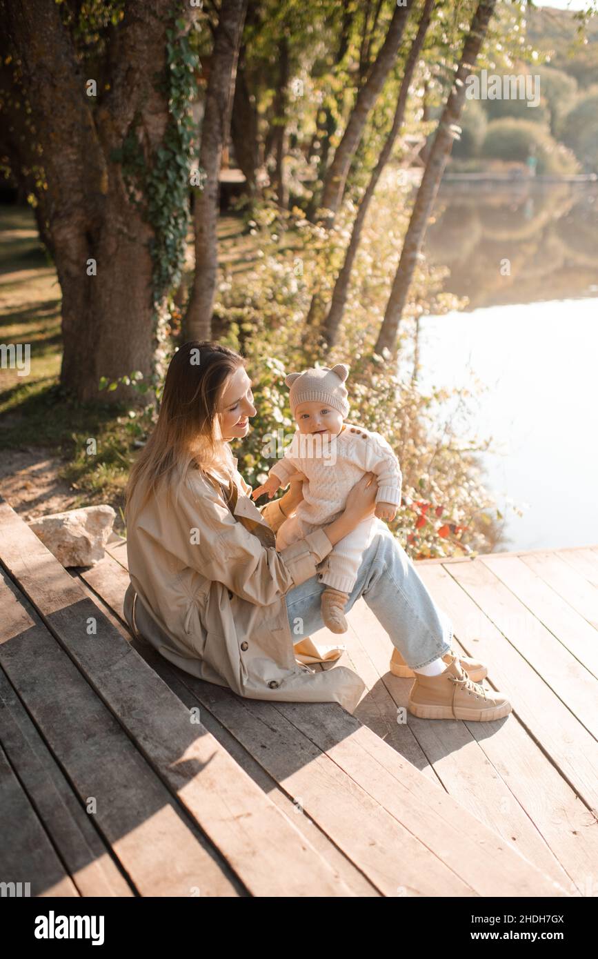 Glücklich lächelnd Mutter tragen Trenchcoat spielen mit Baby junge 1 Jahre  alt tragen Strick Anzug Kleidung sitzen auf hölzernen Pier über See Natur  Hintergrund und s Stockfotografie - Alamy