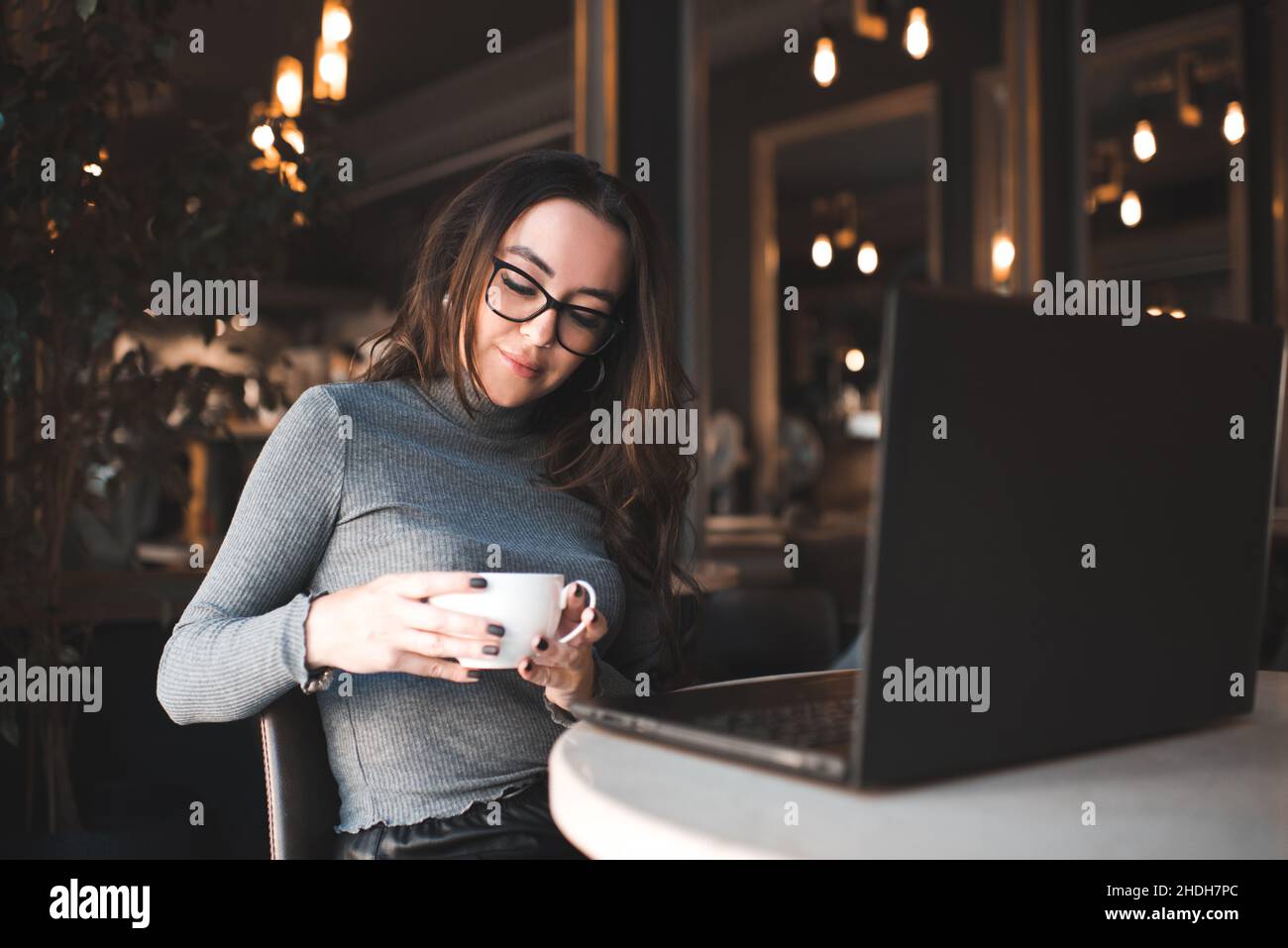 Schöne Brünette Frau mit lockigem Haar sitzt im Café auf Laptop über glühenden Lichtern und gemütliche Atmosphäre arbeiten. Freelancer Mädchen tragen Brille halten Stockfoto