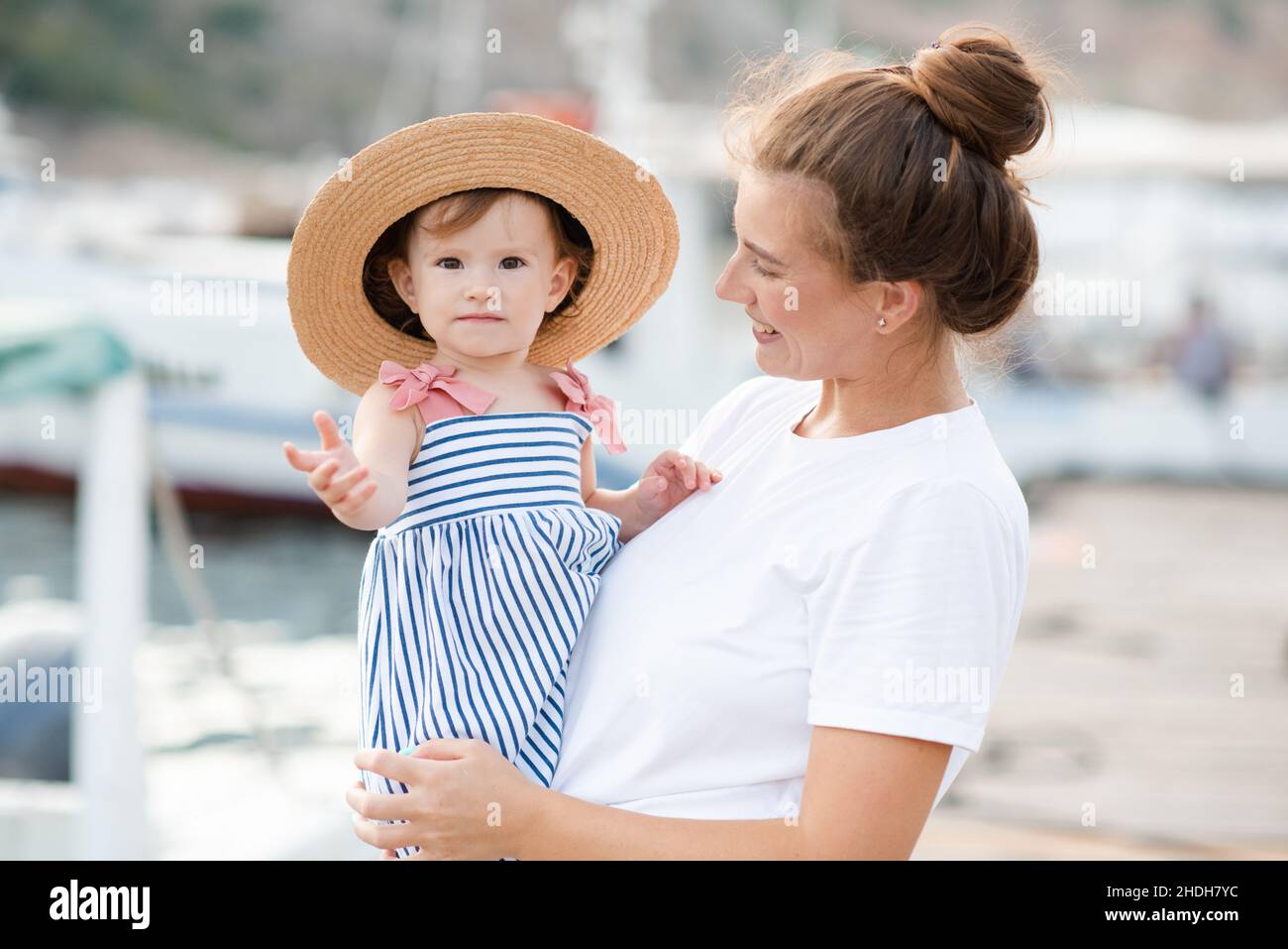 Mutter mit Baby Mädchen 1-2 Jahre alt mit Strohhut über Meer und Boote im Hintergrund aus nächster Nähe. Familienleben. Sommersaison. Kindheit. Stockfoto