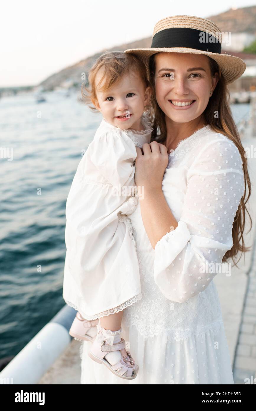 Mutter mit Baby Mädchen 1-2 Jahre alt mit Strohhut über Meer im Hintergrund Nahaufnahme. Familienleben. Sommersaison. Kindheit. Blick auf die Kamera. Stockfoto