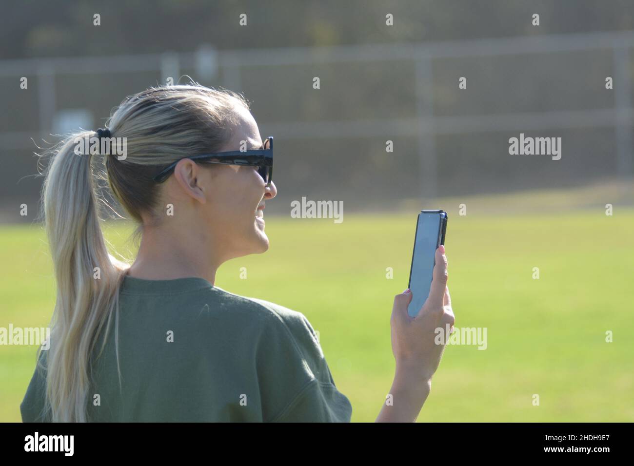 Junge blonde Mutter verwendet Telefon verkaufen, um Töchter Fußballspiel aufzeichnen, mit Sonnenbrille und Pferdeschwanz Stockfoto