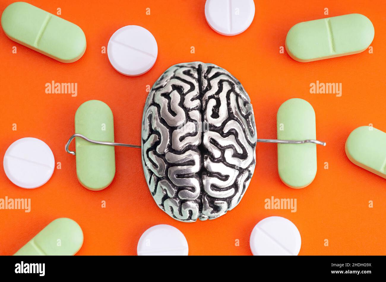 Miniatur-Stahlkopie eines menschlichen Gehirns mit Händen, die Pillen zwischen den verstreuten Drogen auf einem orangefarbenen Hintergrund platziert halten. Schmerzmittel verwenden Störung Konz Stockfoto