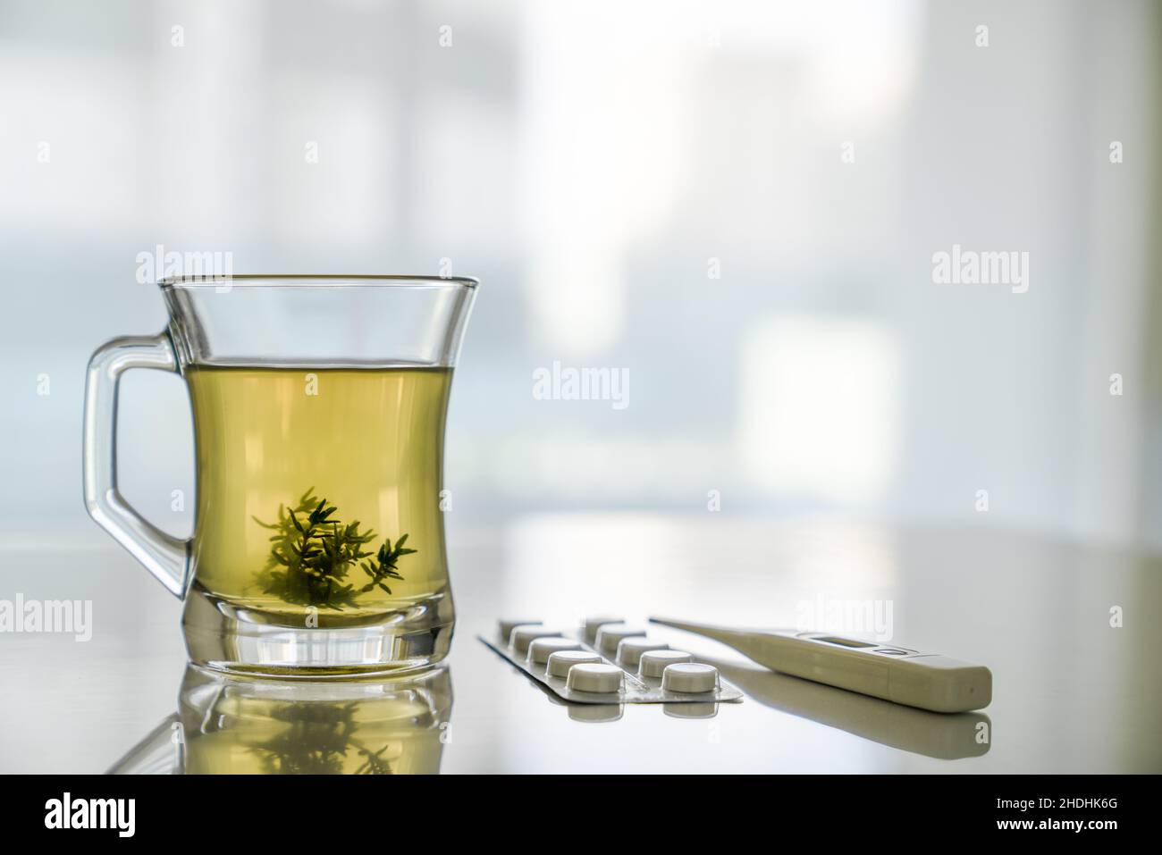 Transparente Glas-Tasse mit Kräutertee und Grasblättern, Thermometer, Tabletten auf weißem Tisch. Konzept der Gesundheitsbehandlung Stockfoto