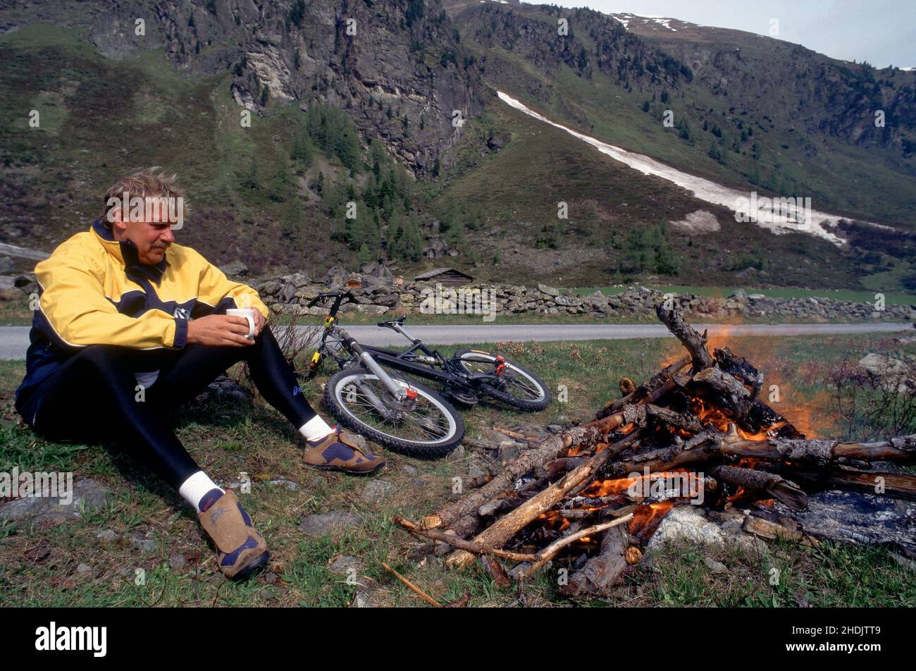 Männlich mit Mountainbike sitzt vor dem Lagerfeuer, um sich aufzuheizen.Abenteuer- und Reisekonzept. Stockfoto