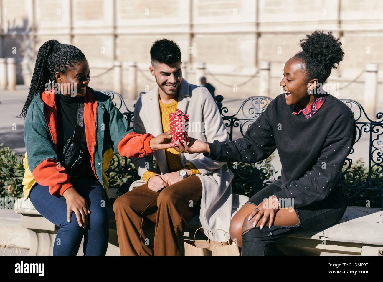 Drei Freunde tauschen Geschenke aus, während sie auf einer Bank auf einem Platz sitzen Stockfoto