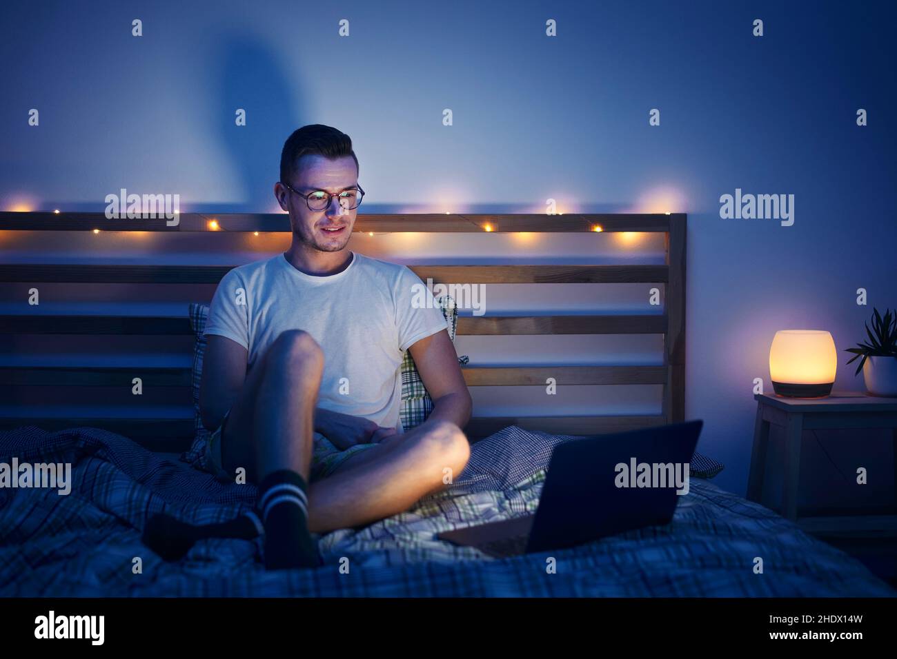 Einsamer Mann mit Brille sitzt im Schlafzimmer auf dem Bett und benutzt nachts einen Laptop. Das Gesicht wird vom Computerdisplay beleuchtet. Stockfoto