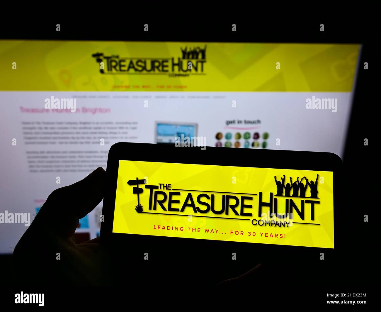 Person, die ein Mobiltelefon mit dem Logo der Treasure Hunt Company (Brighton) auf dem Bildschirm vor der Unternehmenswebseite hält. Konzentrieren Sie sich auf die Telefonanzeige. Stockfoto