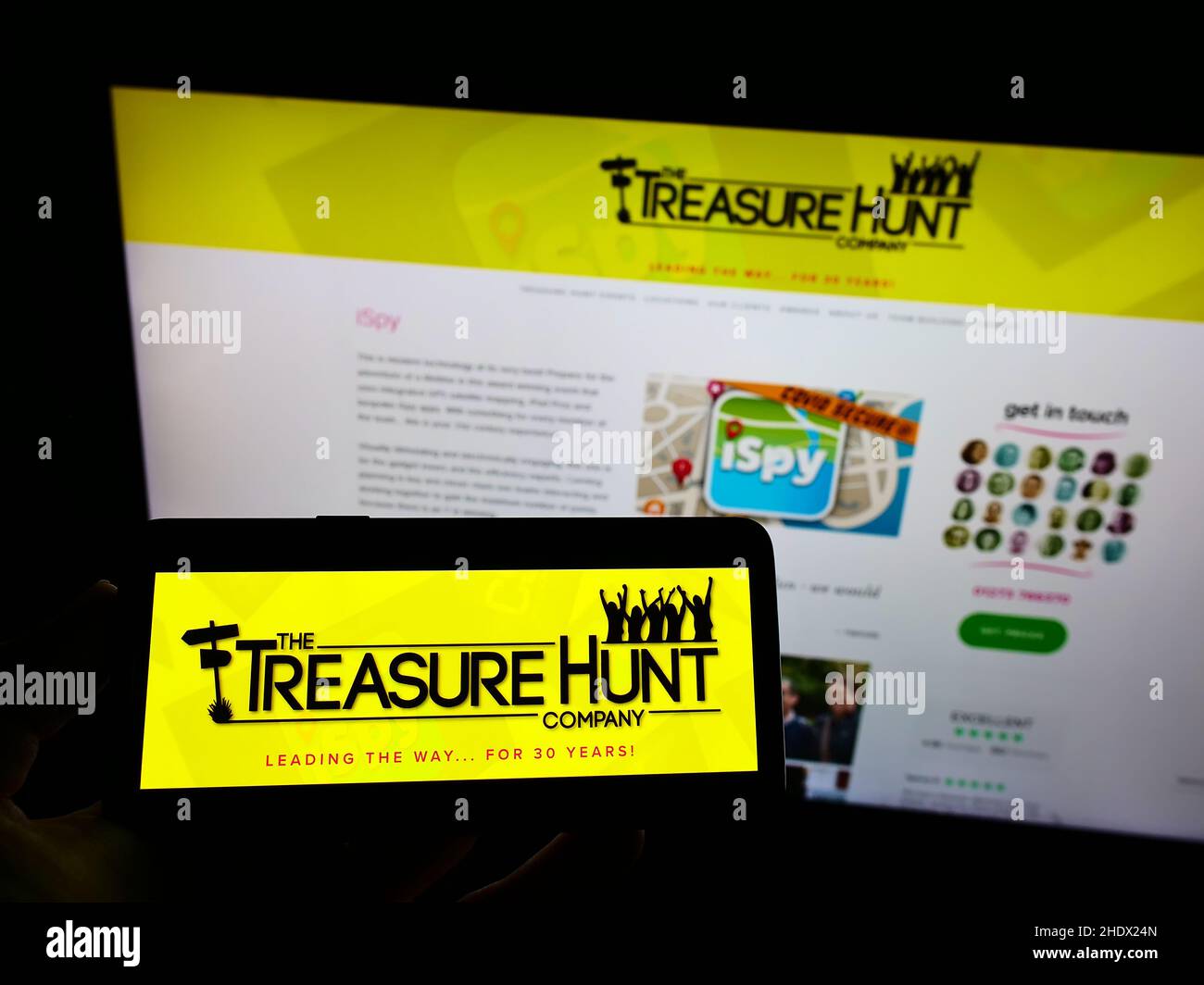Person mit Mobiltelefon und Logo der Treasure Hunt Company (Brighton) auf dem Bildschirm vor der Business-Webseite. Konzentrieren Sie sich auf die Telefonanzeige. Stockfoto