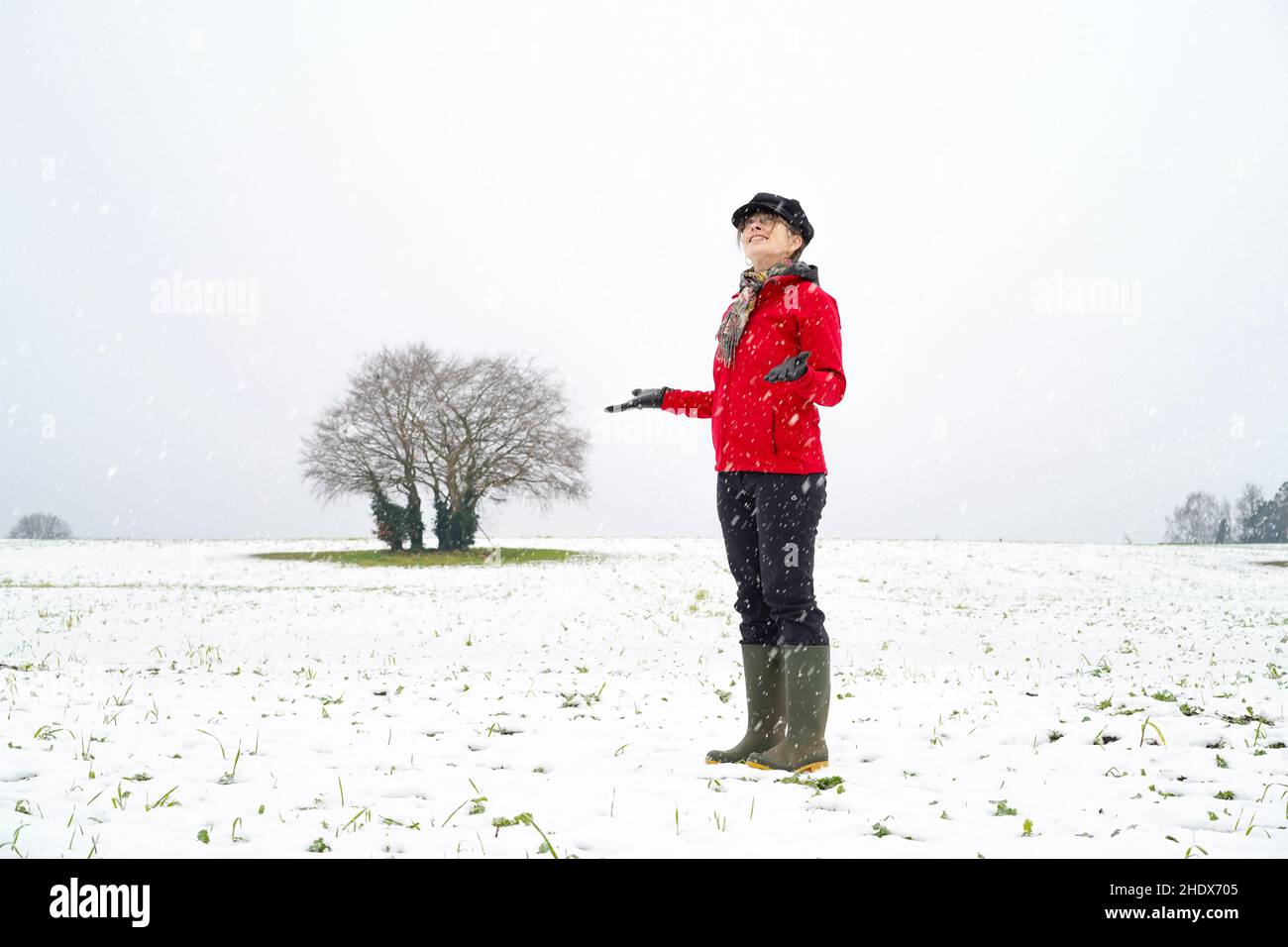 Die junge lächelnde Frau in knallroter Jacke und trendigem Hut steht isoliert auf Feldern auf dem Land, hält die Hände heraus und blickt auf fallenden Winterschnee. Stockfoto