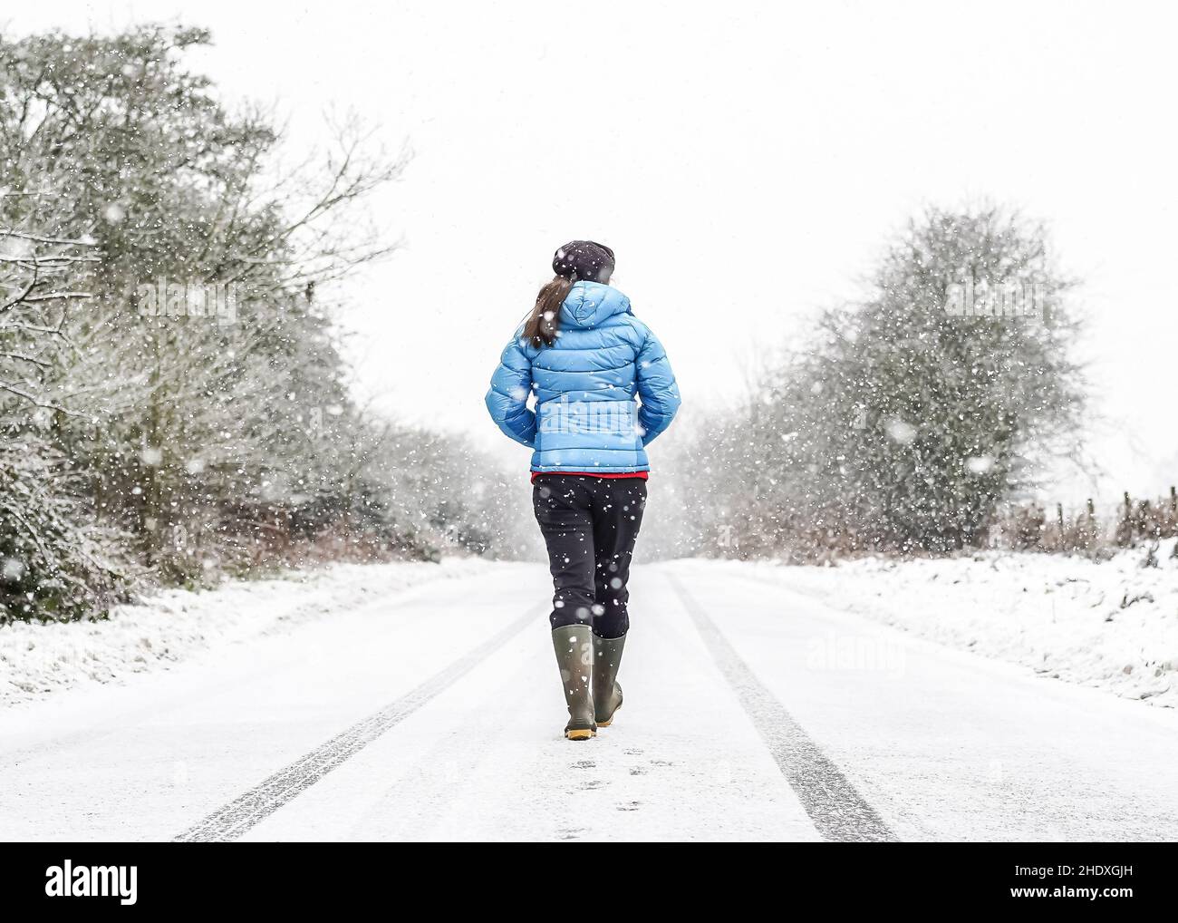 Rückansicht einer jungen Frau in Wintermantel und Gummistiefeln, isoliert im Freien auf der britischen Landschaft, die mitten auf einer verlassenen Landstraße im fallenden Schnee läuft. Stockfoto
