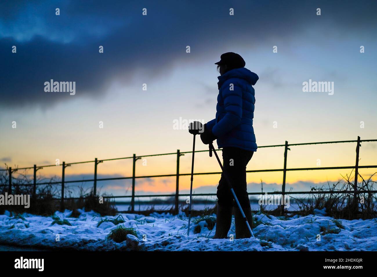 Nahansicht der Frau (mit Hut und Wintermantel) in einer isolierten Silhouette im Freien, die in der Dämmerung im Schnee läuft, mit Sonnenuntergang am Himmel. Stockfoto