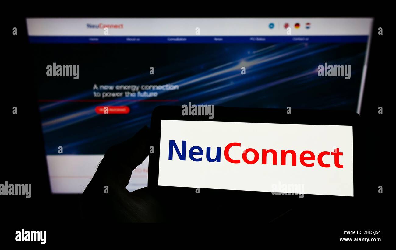 Person, die das Smartphone mit dem Logo des Kraftübertragungsprojekts NeuConnect Interconnector auf dem Bildschirm vor der Website hält. Konzentrieren Sie sich auf die Telefonanzeige. Stockfoto