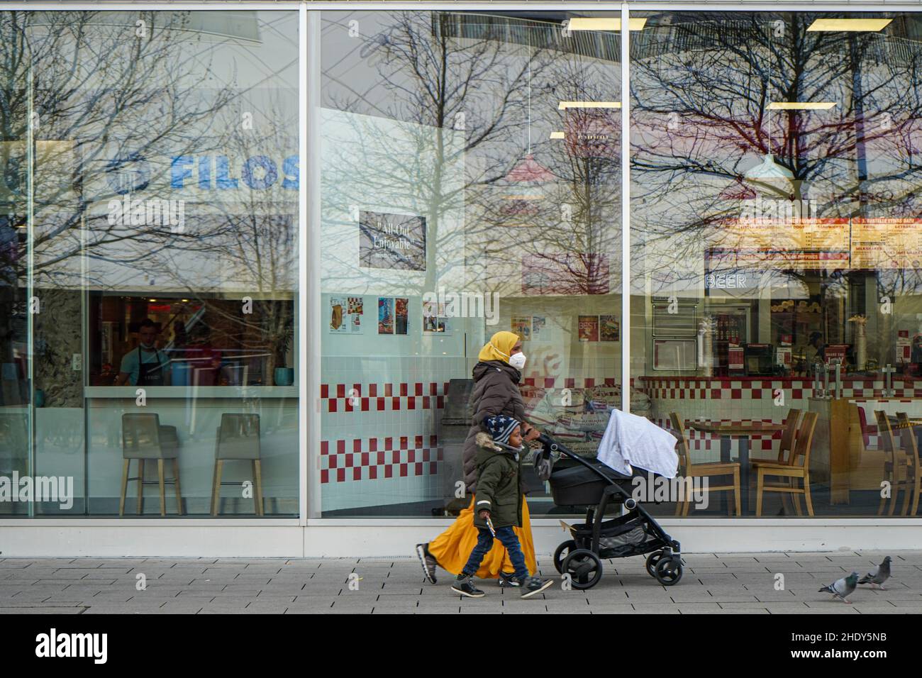 Eine Frau, die eine Covid-Maske trägt, einen Pram vor sich schiebt, ihr kleiner Sohn, der neben ihr läuft, passiert die Fenster eines Einkaufszentrums. Stockfoto