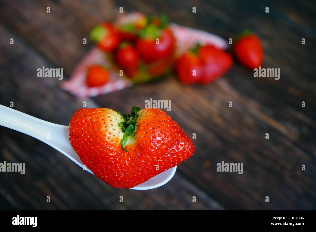 Eine einzigartige herzförmige Bio-Erdbeere auf einem weißen Löffel. Erdbeeren mit rot-weiß karierter Tischdecke auf einem rustikalen Holztisch im Hintergrund. Stockfoto