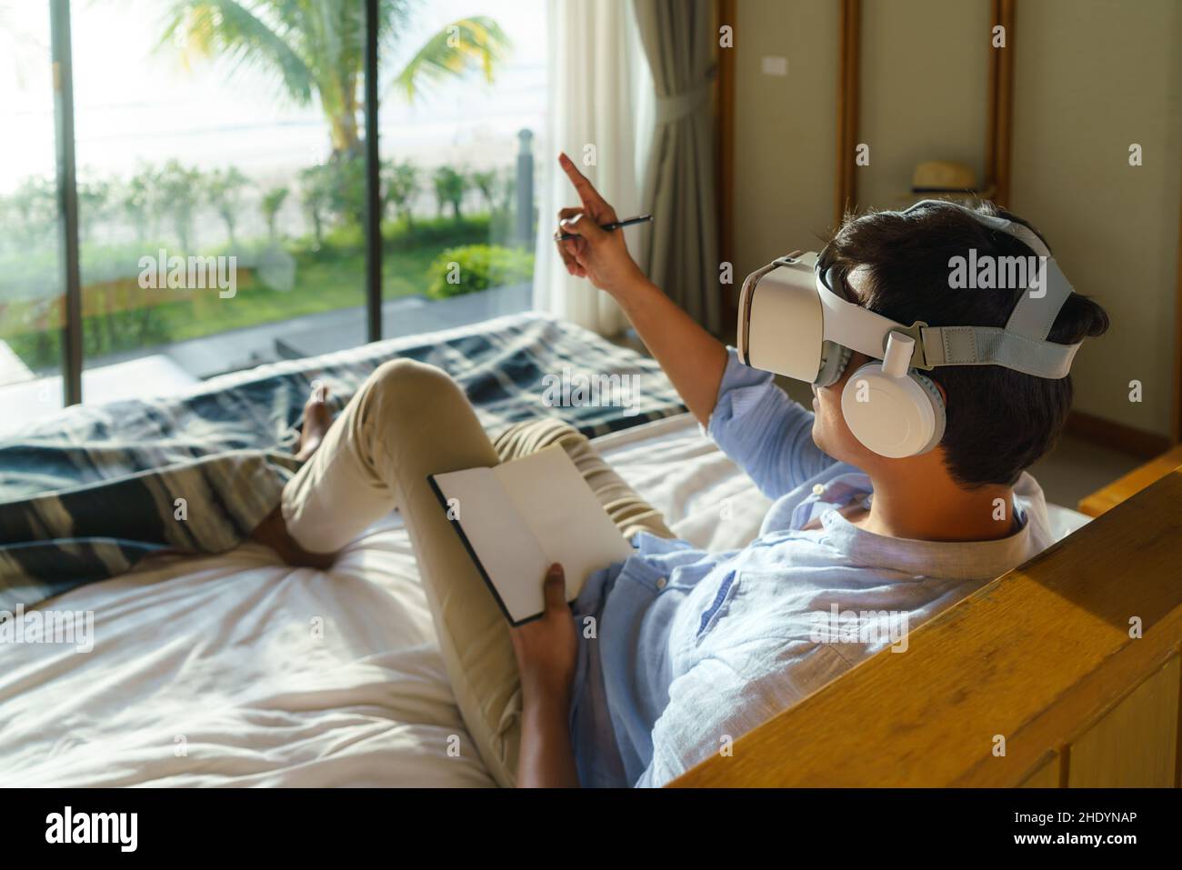 Junger asiatischer Mann, der während seines Sommerurlaubs auf dem Bett liegt und eine Virtual-Reality-Brille für Geschäftstreffen verwendet. Stockfoto