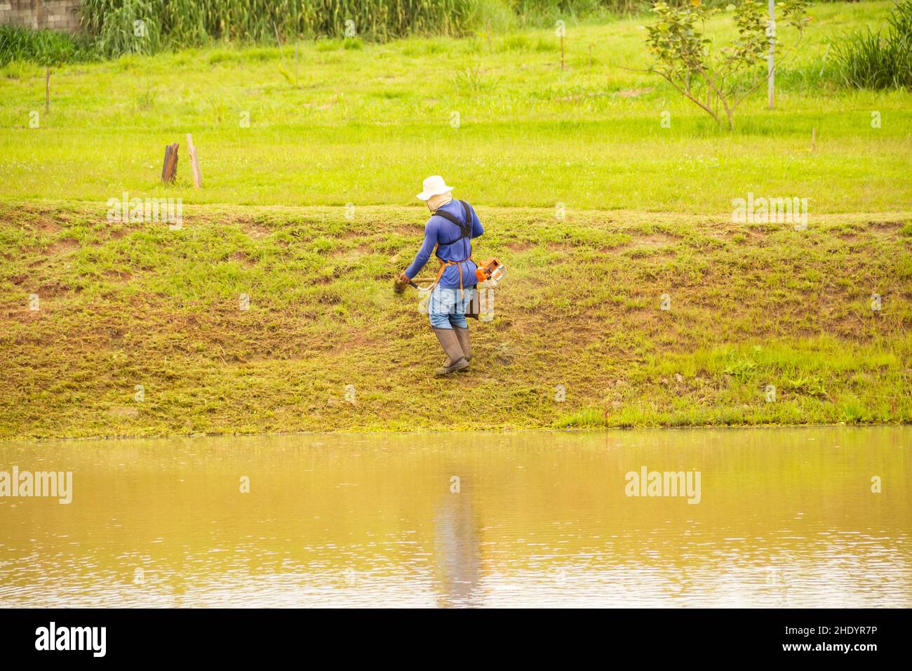 Goiânia, Goias, Brasilien – 05. Januar 2022: Ein Arbeiter, der mit entsprechender Ausrüstung Gras am Seeufer schneidet. Stockfoto