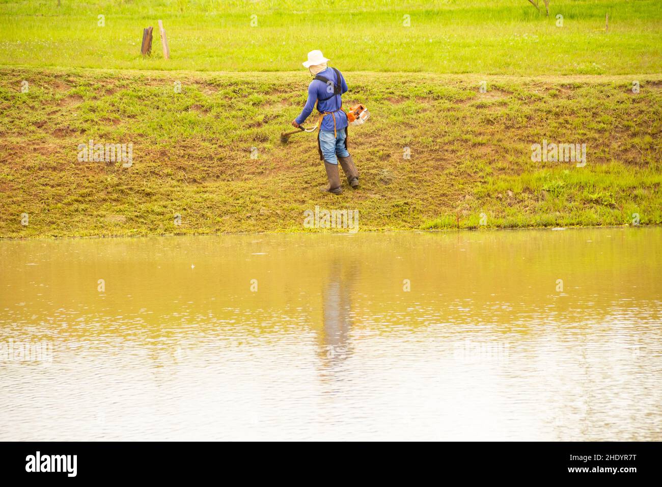 Goiânia, Goias, Brasilien – 05. Januar 2022: Ein Arbeiter, der mit entsprechender Ausrüstung Gras am Seeufer schneidet. Stockfoto