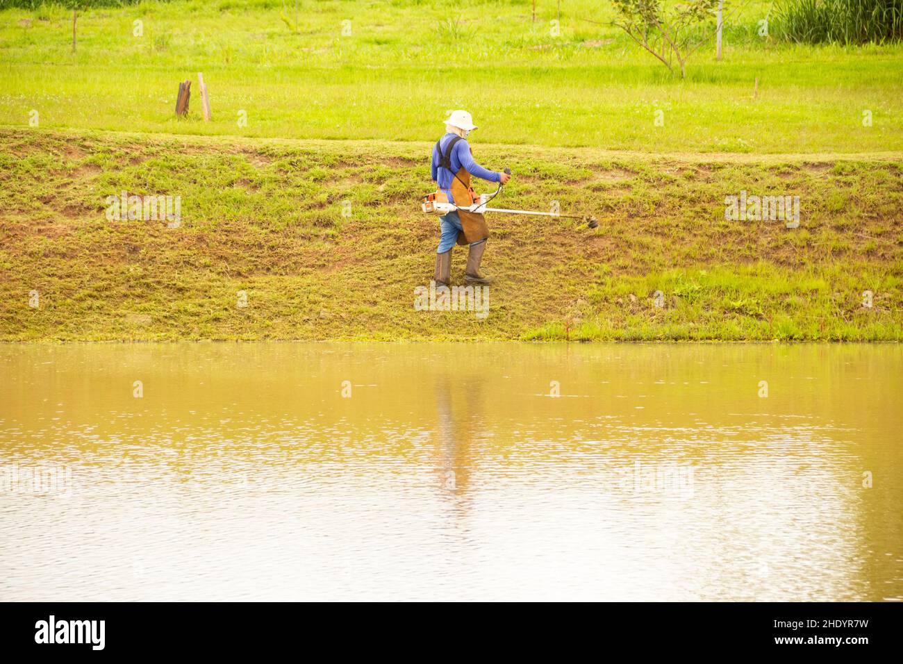 Goiânia, Goias, Brasilien – 05. Januar 2022: Ein Arbeiter, der mit entsprechender Ausrüstung Gras am Seeufer schneidet. Stockfoto