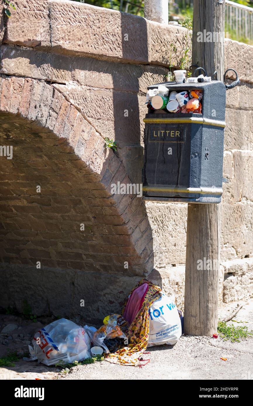 Der Abfalleimer auf einem Pfosten, der mit Abfall überfüllt ist, und die Beuteltaschen auf dem Boden darunter Stockfoto