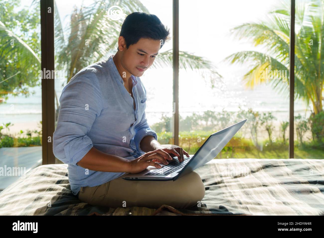 Ein junger asiatischer Geschäftsmann sitzt im Bett und arbeitet während eines Sommerurlaubs im Zimmer des Resorts in der Nähe des Meeres mit einem Laptop. Stockfoto Ein junger asiatischer Geschäftsmann sitzt im Bett und arbeitet während eines Sommerurlaubs im Zimmer des Resorts in der Nähe des Meeres mit einem Laptop. Stockfoto