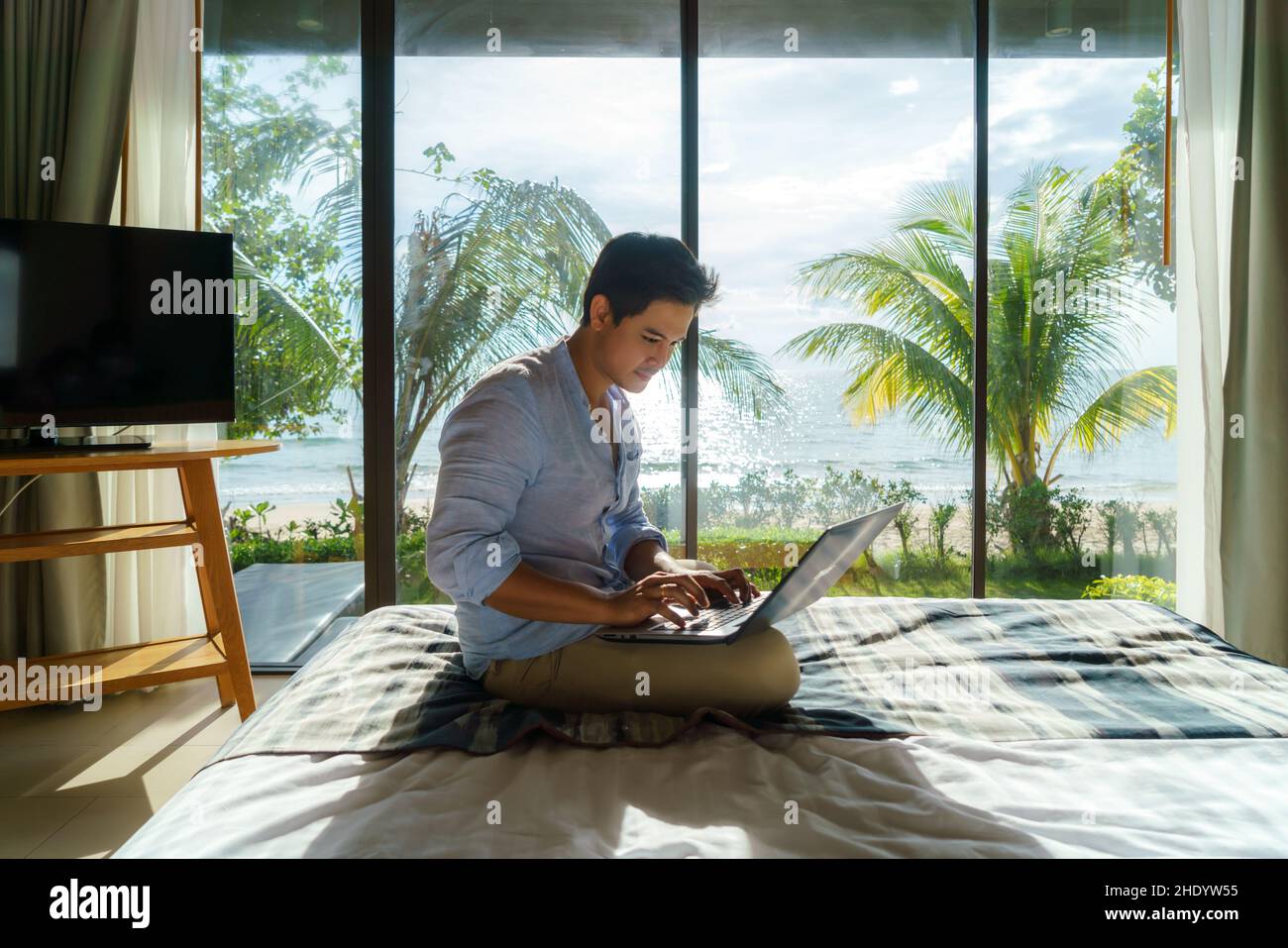 Ein junger asiatischer Geschäftsmann sitzt im Bett und arbeitet während eines Sommerurlaubs im Zimmer des Resorts in der Nähe des Meeres mit einem Laptop. Stockfoto Ein junger asiatischer Geschäftsmann sitzt im Bett und arbeitet während eines Sommerurlaubs im Zimmer des Resorts in der Nähe des Meeres mit einem Laptop. Stockfoto