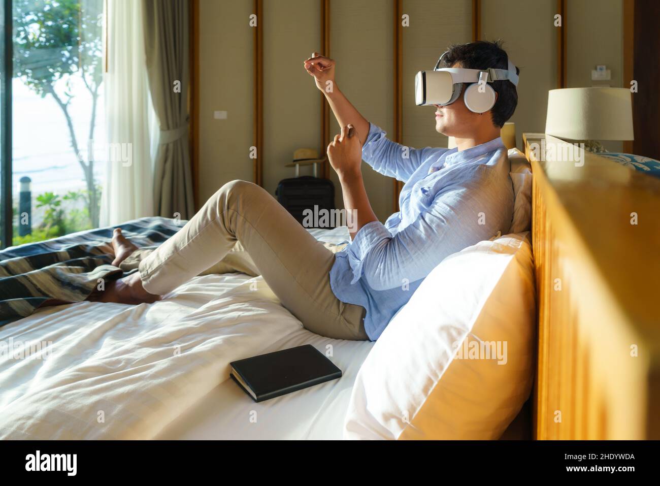 Junger asiatischer Mann, der während seines Sommerurlaubs auf dem Bett liegt und eine Virtual-Reality-Brille für Geschäftstreffen verwendet. Stockfoto