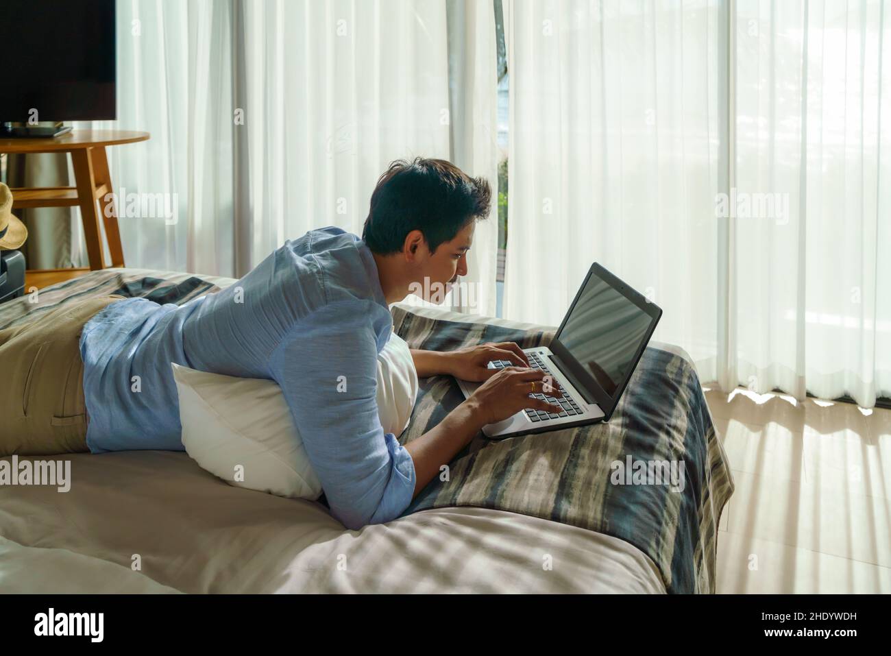 Ein junger asiatischer Geschäftsmann liegt im Bett und arbeitet während eines Sommerurlaubs im Zimmer des Resorts in der Nähe des Meeres mit einem Laptop. Stockfoto Ein junger asiatischer Geschäftsmann liegt im Bett und arbeitet während eines Sommerurlaubs im Zimmer des Resorts in der Nähe des Meeres mit einem Laptop. Stockfoto