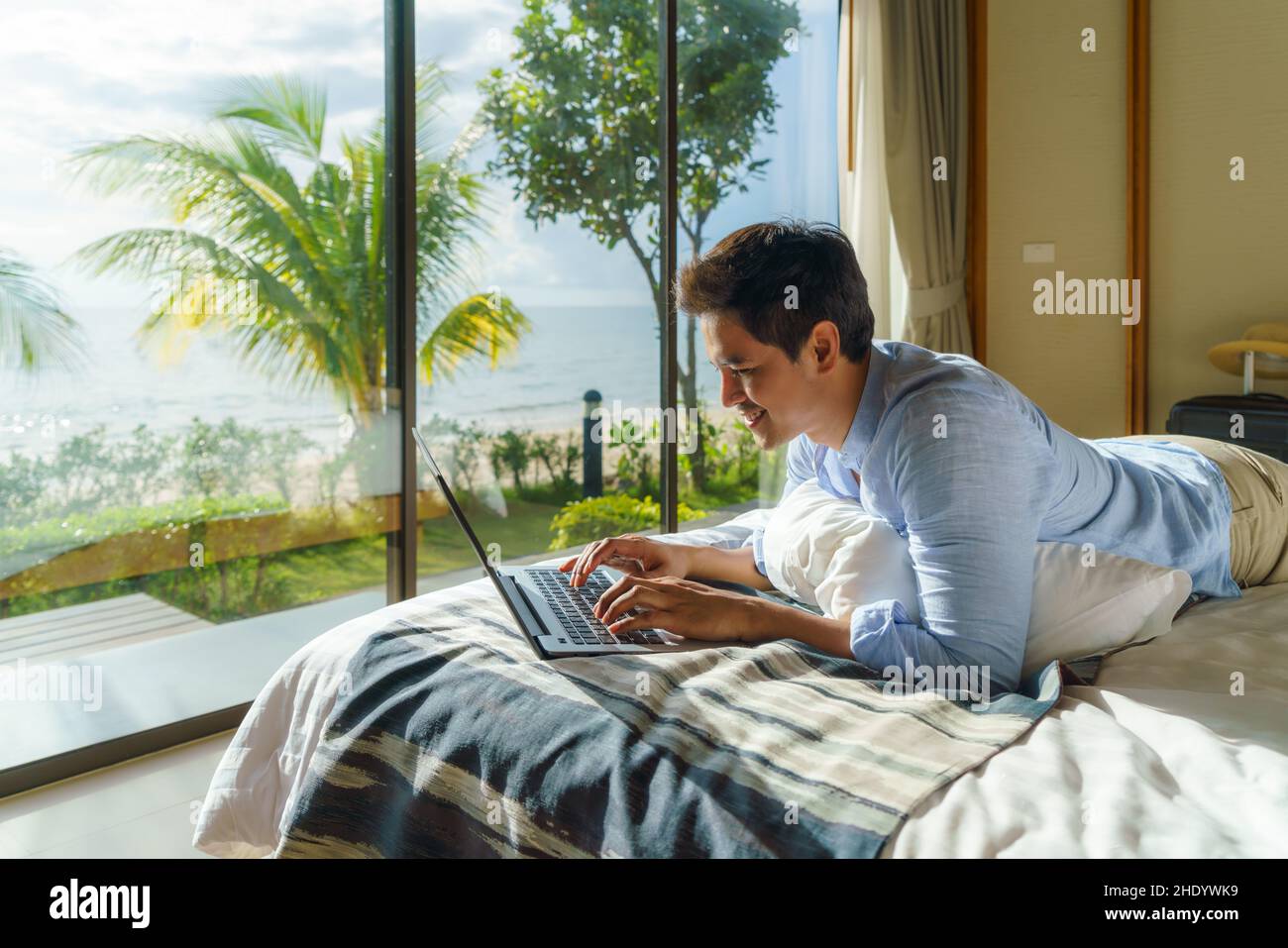 Ein junger asiatischer Geschäftsmann liegt im Bett und arbeitet während eines Sommerurlaubs im Zimmer des Resorts in der Nähe des Meeres mit einem Laptop. Stockfoto Ein junger asiatischer Geschäftsmann liegt im Bett und arbeitet während eines Sommerurlaubs im Zimmer des Resorts in der Nähe des Meeres mit einem Laptop. Stockfoto