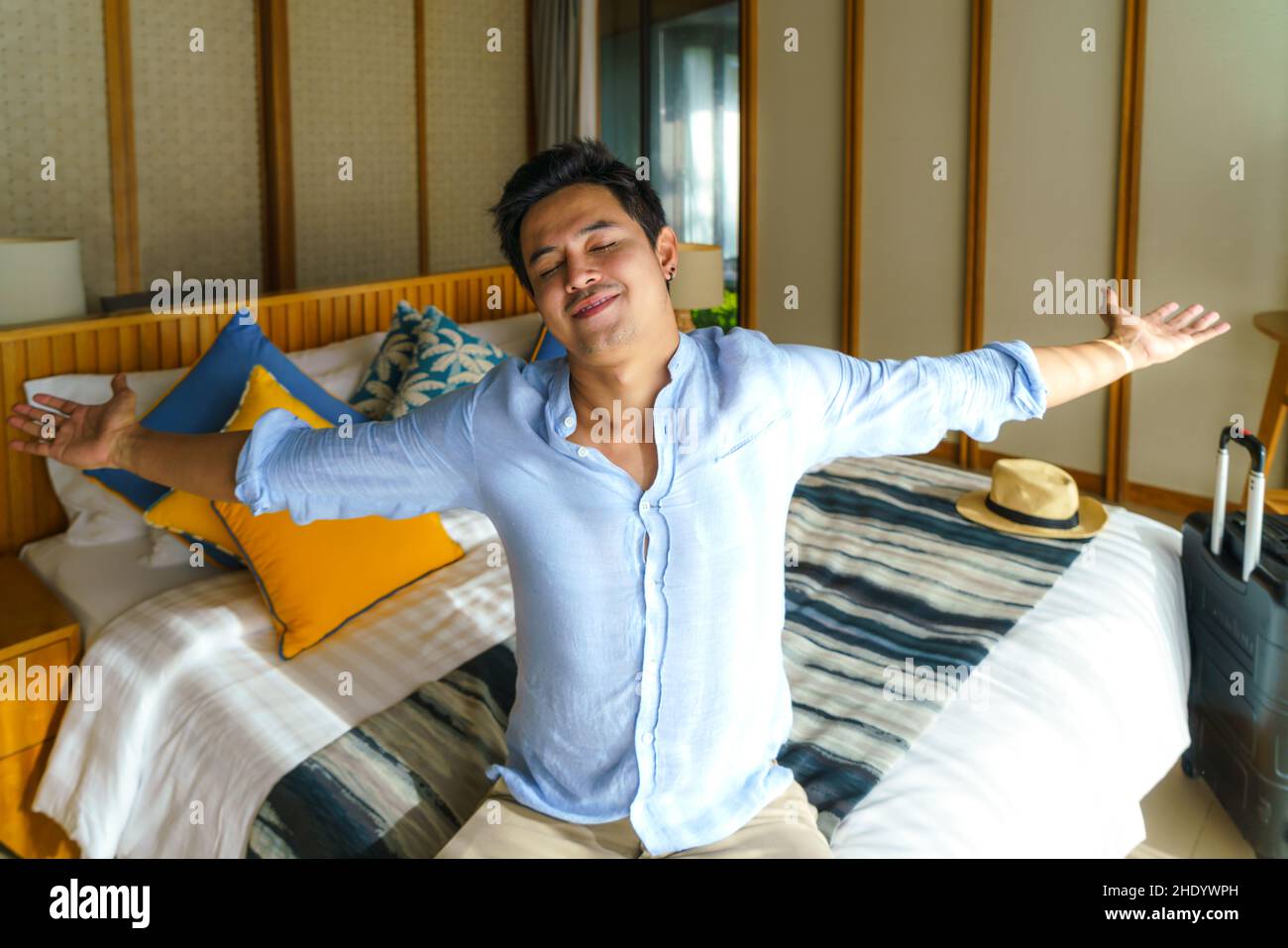 Ein asiatischer Mann mit Koffer legte sich nach seiner Ankunft während seiner Sommerurlaubsreise in seinem Zimmer im Resort auf ein Bett. Stockfoto