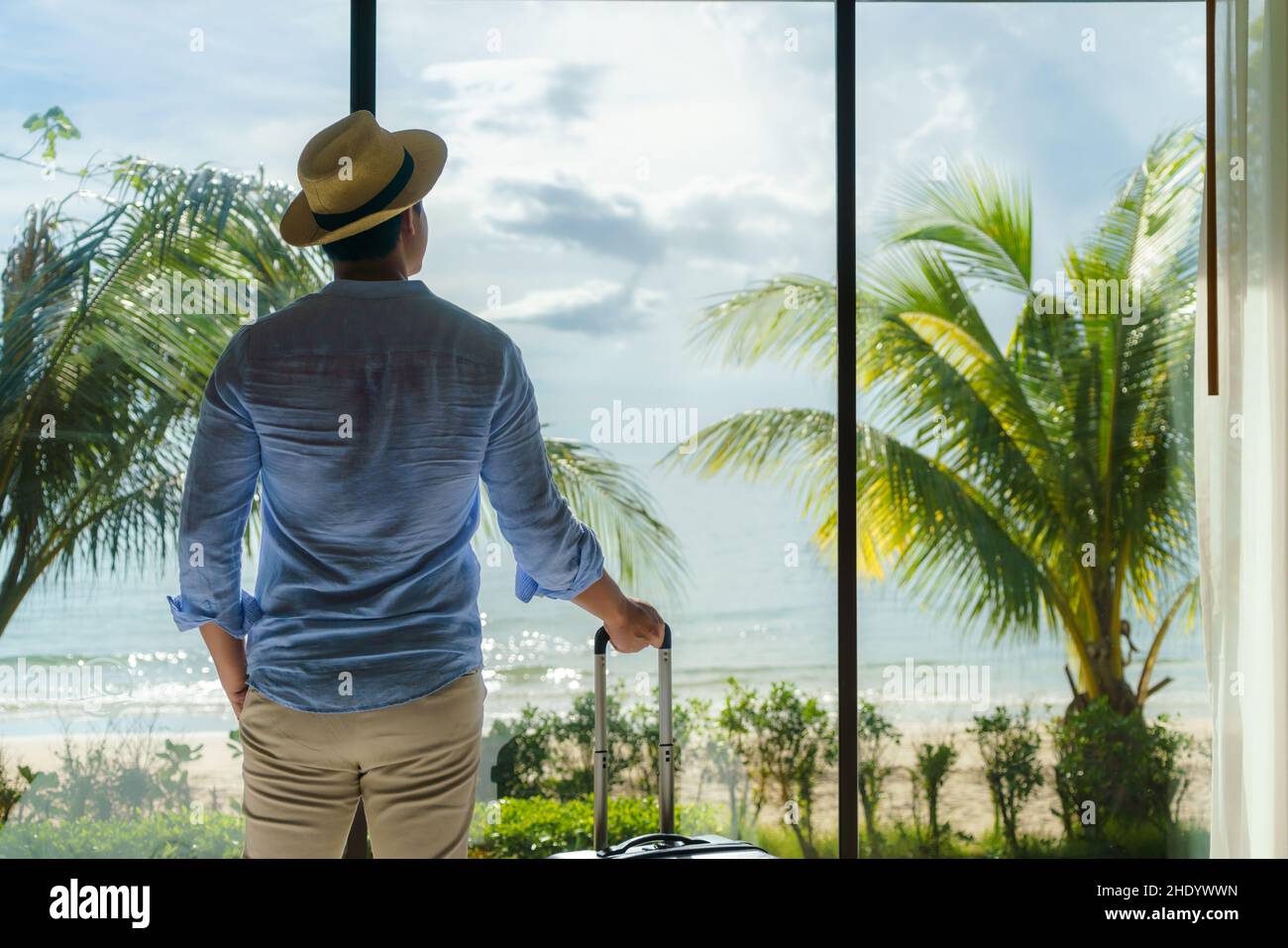 Ein asiatischer Mann mit Koffer steht neben dem Fenster seines Zimmers und blickt während seiner Sommerurlaubsreise auf den wunderschönen Meerblick. Stockfoto Ein asiatischer Mann mit Koffer steht neben dem Fenster seines Zimmers und blickt während seiner Sommerurlaubsreise auf den wunderschönen Meerblick. Stockfoto