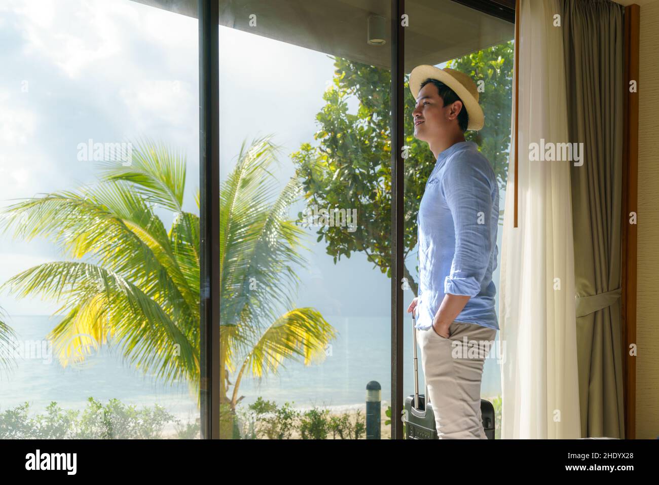 Ein asiatischer Mann mit Koffer steht neben dem Fenster seines Zimmers und blickt während seiner Sommerurlaubsreise auf den wunderschönen Meerblick. Stockfoto Ein asiatischer Mann mit Koffer steht neben dem Fenster seines Zimmers und blickt während seiner Sommerurlaubsreise auf den wunderschönen Meerblick. Stockfoto