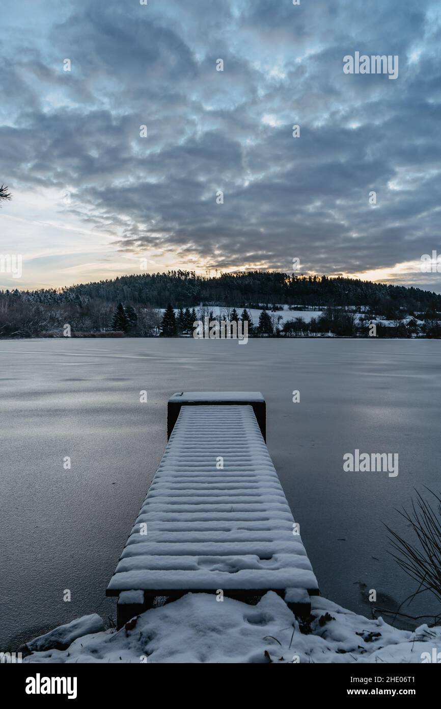 Hölzerner Pier am See mit frischem Schnee.Winterteich mit kleinem Steg bei Sonnenaufgang, Wald im Hintergrund.Frostige ruhige Landschaft. Weiße Winterlandschaft verschneit Stockfoto