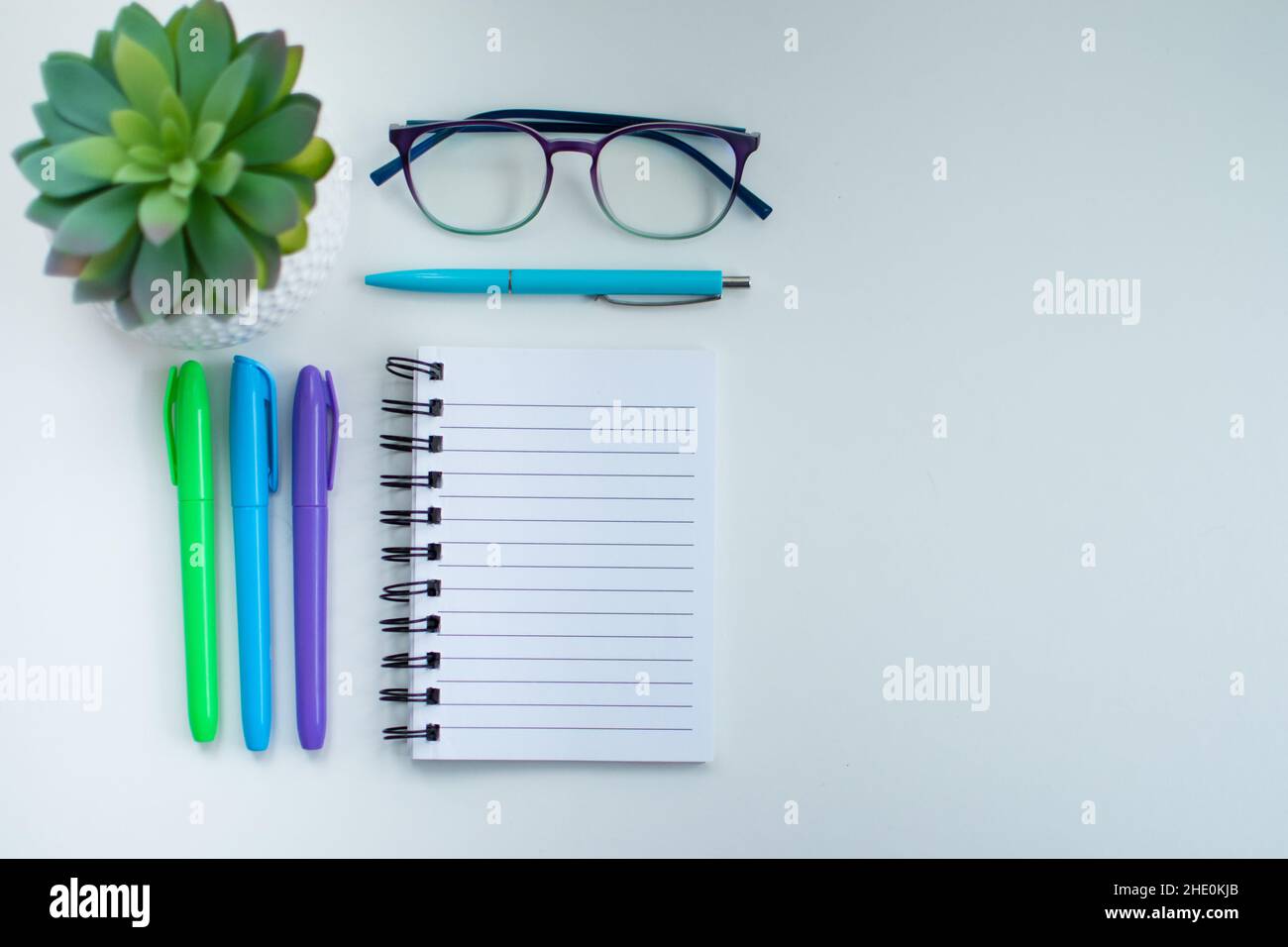 Hoher Winkel eines weißen Schreibtisches mit bunten Schreibgeräte darauf und einer eingetopften grünen Pflanze, einer Brille und einem leeren Notizbuch Stockfoto