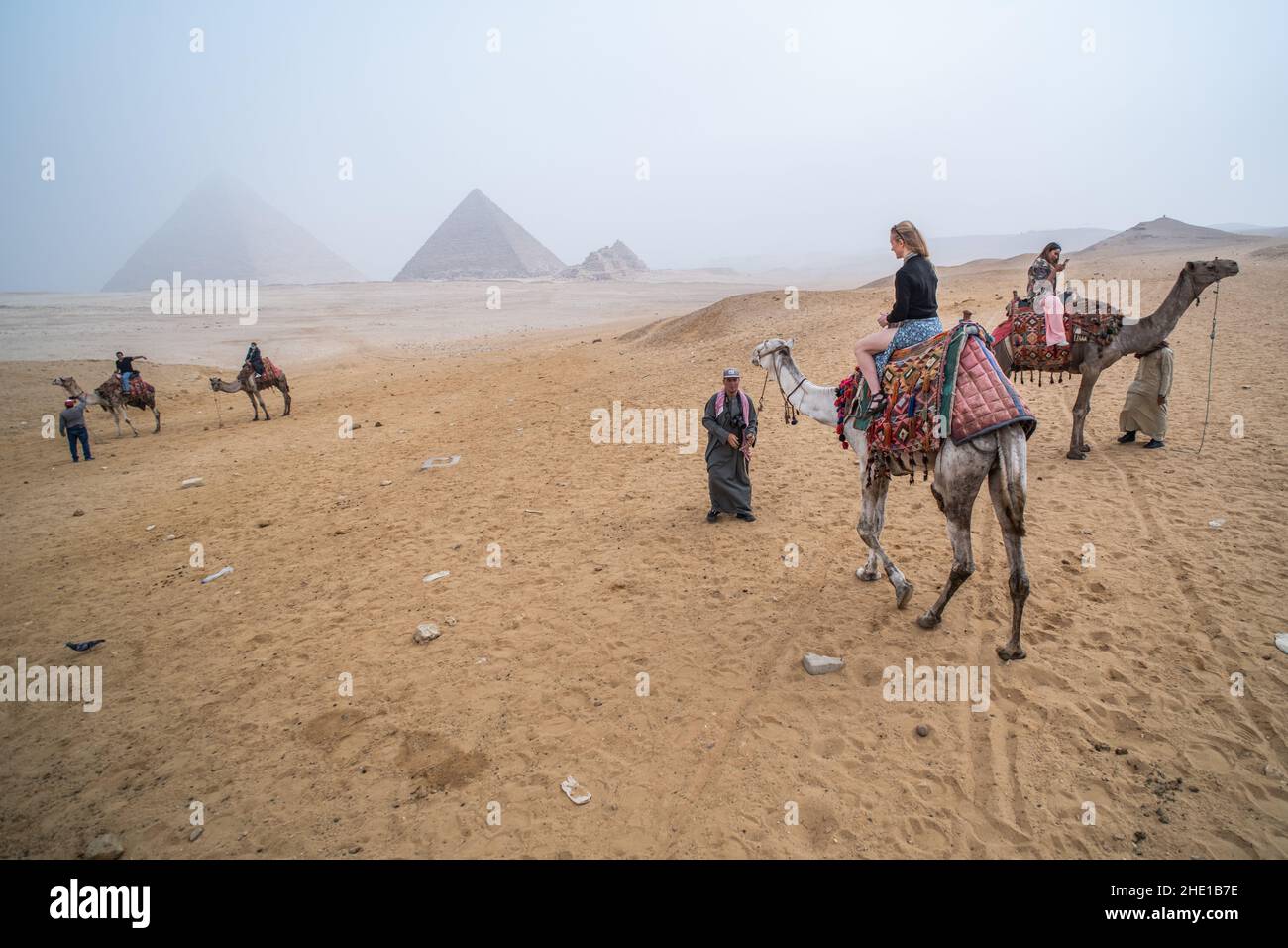 Eine alleinreisende Frau auf einem Kamelritt in der Wüste mit den Pyramiden von Gizeh, die durch starken Smog sichtbar sind. Stockfoto