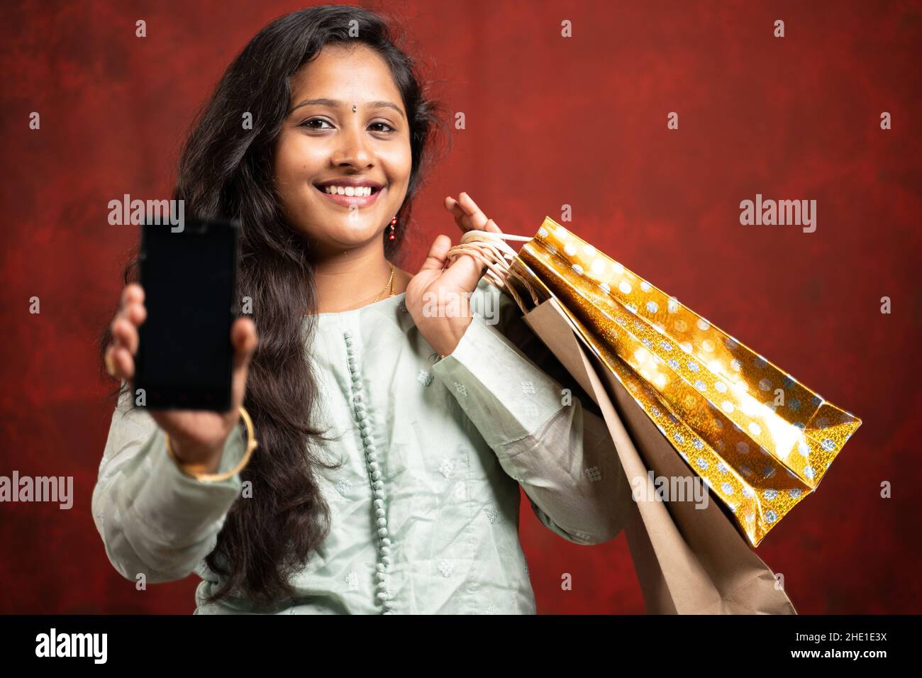 Glücklich lächelndes indisches Mädchen mit Einkaufstaschen, die Handy zeigen, indem sie die Kamera betrachten - ein Konkpet von Online-E-Commerce-Shopping und Festivalverkäufen. Stockfoto