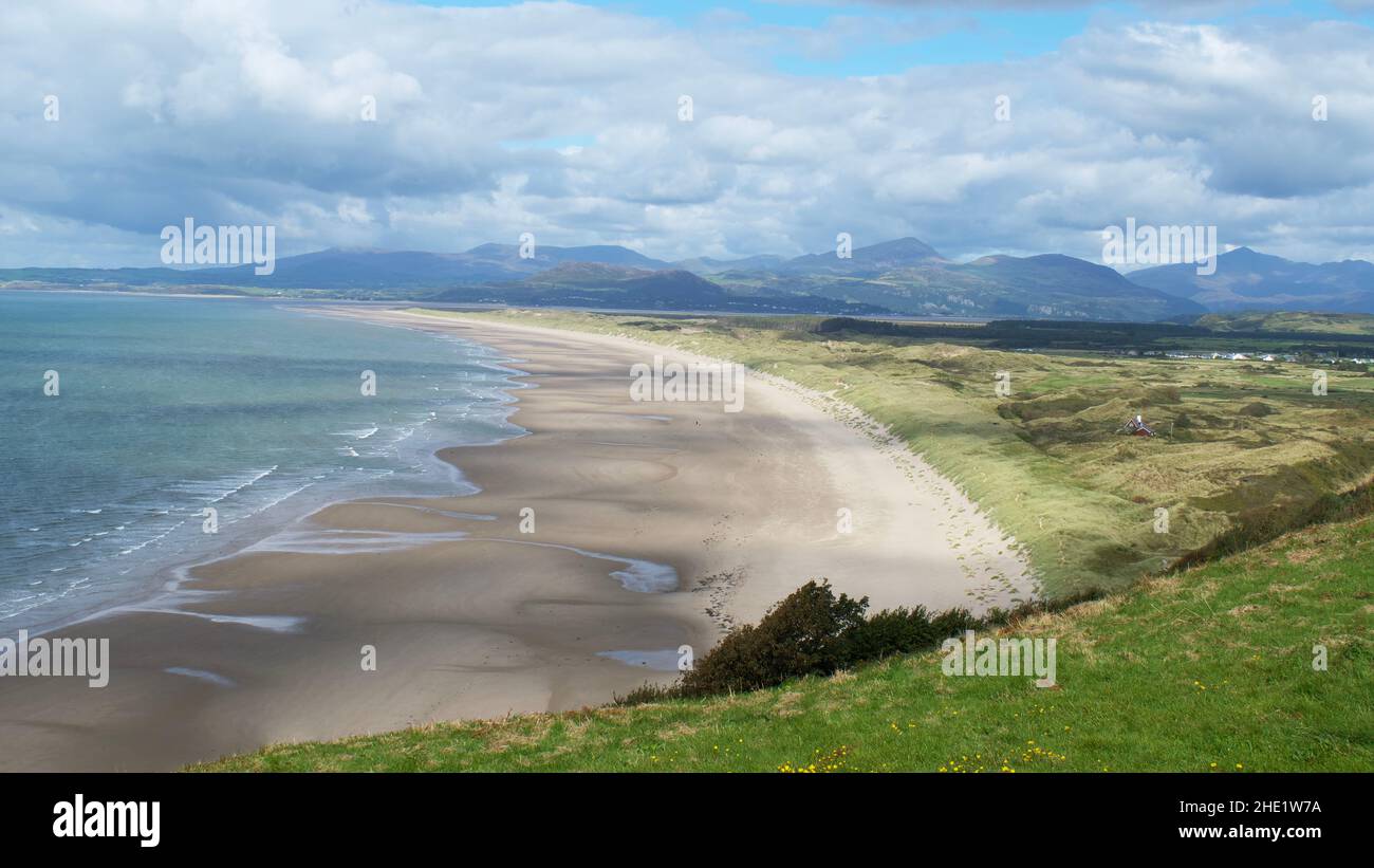 Weitläufiger sandiger Harlech Beach mit sanften Wellen und grasbewachsenen Dünen, umgeben von fernen Bergen unter teilweise bewölktem Himmel, Gwynedd, Wales, Großbritannien. Stockfoto