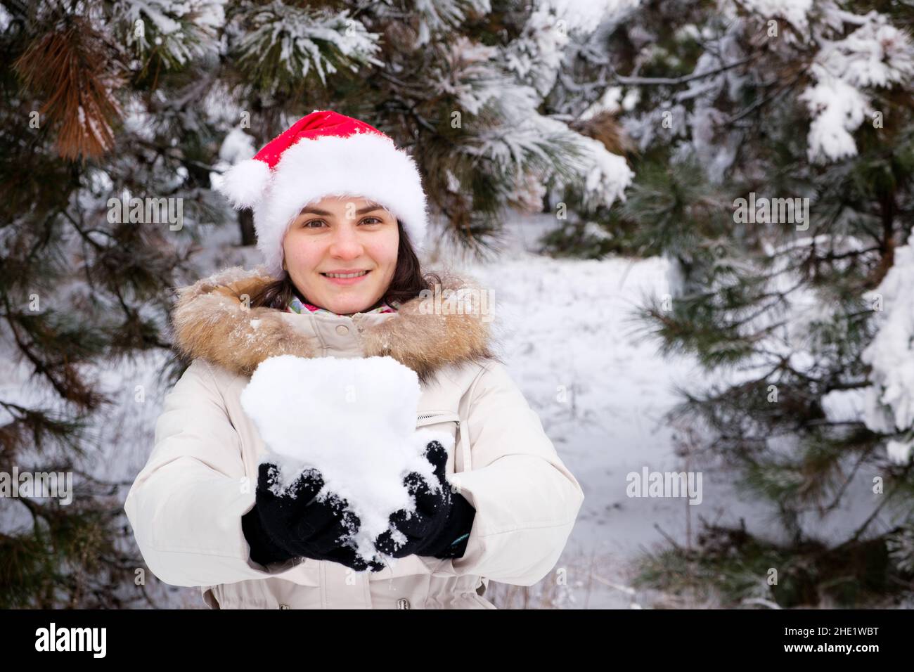 Eine Frau im Weihnachtsmann-Hut hält einen Schneeball in ihren Händen in einem schneebedeckten Winterwald, ein Ort für Text Stockfoto