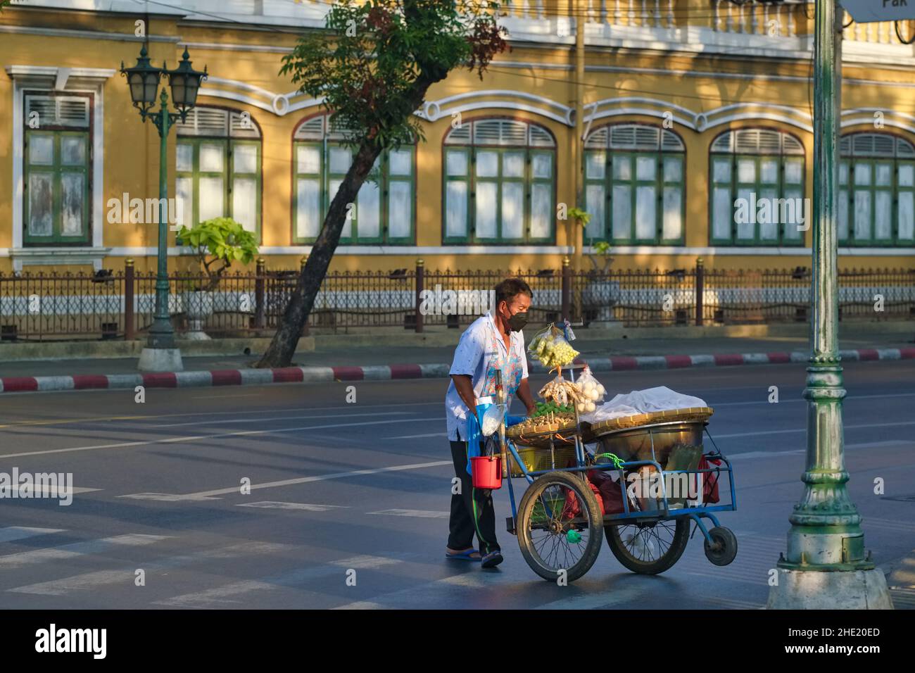 Ein Lebensmittelhändler mit seinem Trolley wird vom Licht des frühen Morgens erleuchtet und fährt an der Suankularb Wittayalai School im Kolonialstil, Tripetch Rd., Bangkok, Thailand, vorbei Stockfoto