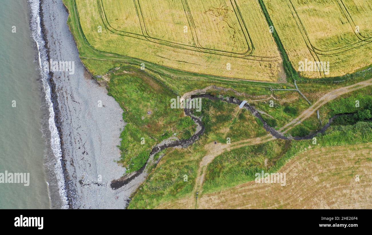 Luftbild eines gewundenen Baches, der zum Meer fließt, können Sie Felder, Strand und Meer sehen. Der Küstenweg führt über den Bach über eine große Schieferplatte. Stockfoto