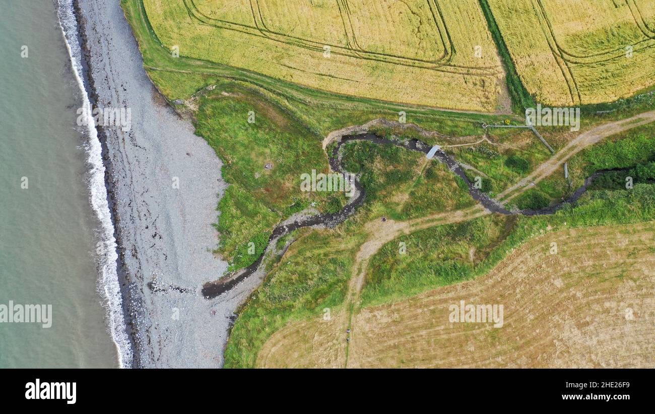 Luftbild eines gewundenen Baches, der zum Meer fließt, können Sie Felder, Strand und Meer sehen. Der Küstenweg führt über den Bach über eine große Schieferplatte. Stockfoto