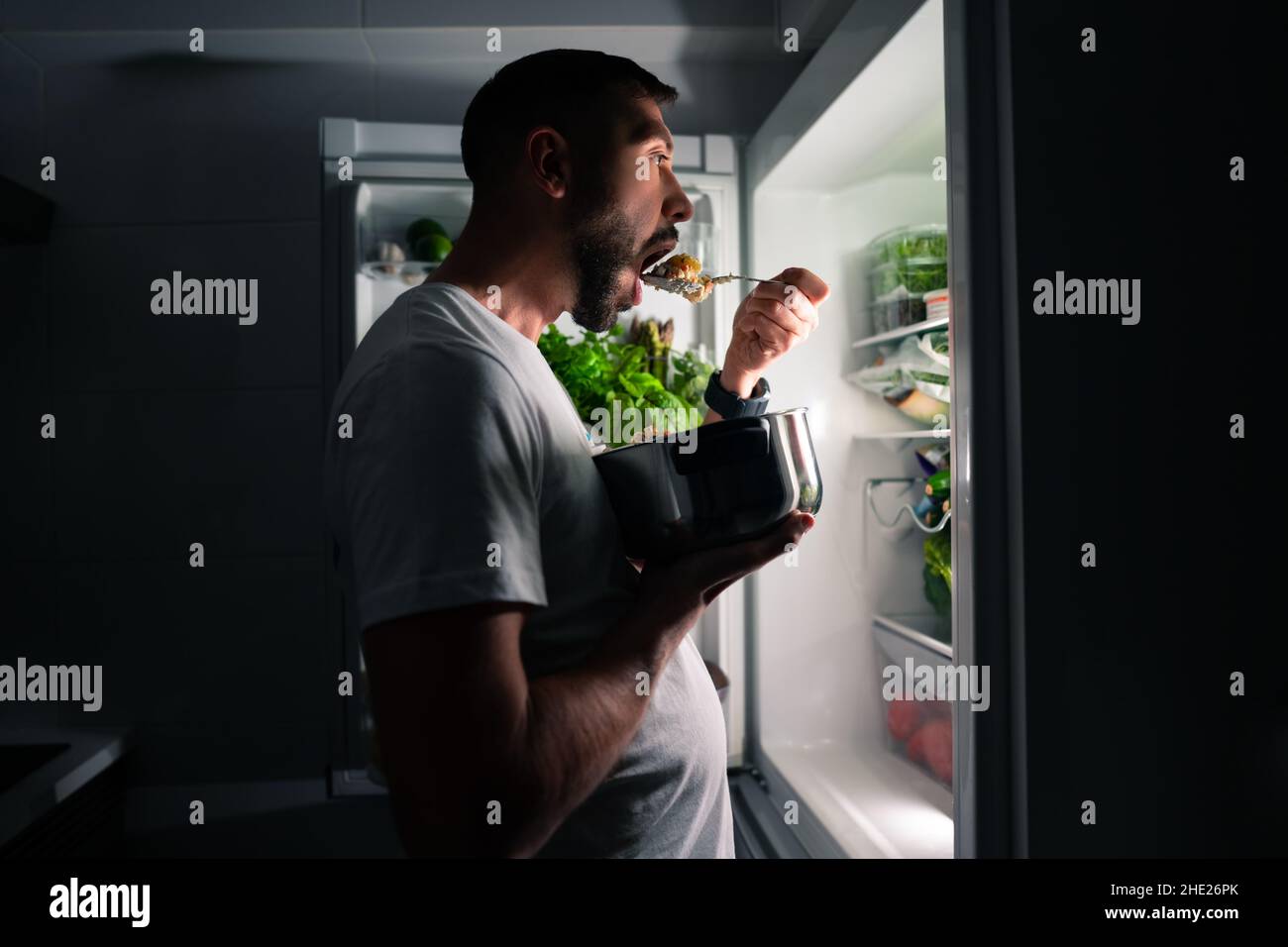 Junger hungriger Mann, der nachts Essen isst und in einen offenen Kühlschrank schaut. Mann, der Mitternachtssnack aus dem Kühlschrank nahm Stockfoto