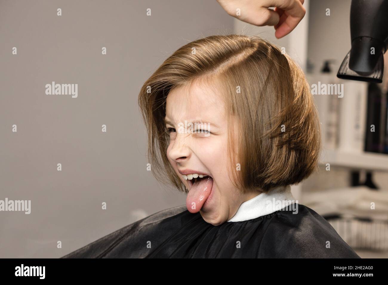 Expressive kurzhaarige kleine Mädchen macht Grimassen und zeigt Zunge in Friseurladen in Prozess der Trocknung der Haare im Schönheitssalon. Friseursalon. Friseurdienste. Schönheitskonzept. Stockfoto
