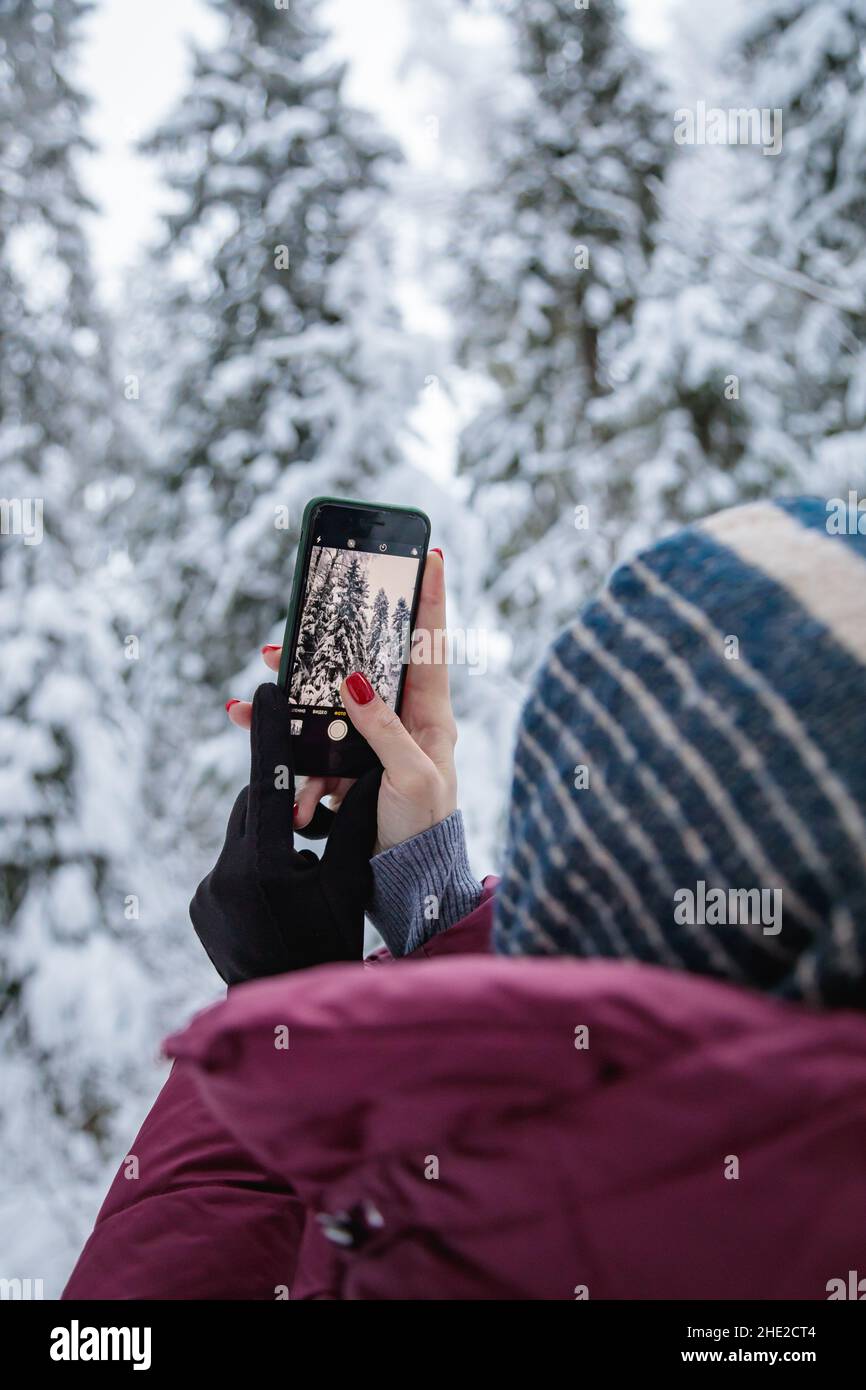 Ein Mädchen in einer burgunderroten Daunenjacke fotografiert auf einem Mobiltelefon einen schneebedeckten Wald. Stockfoto