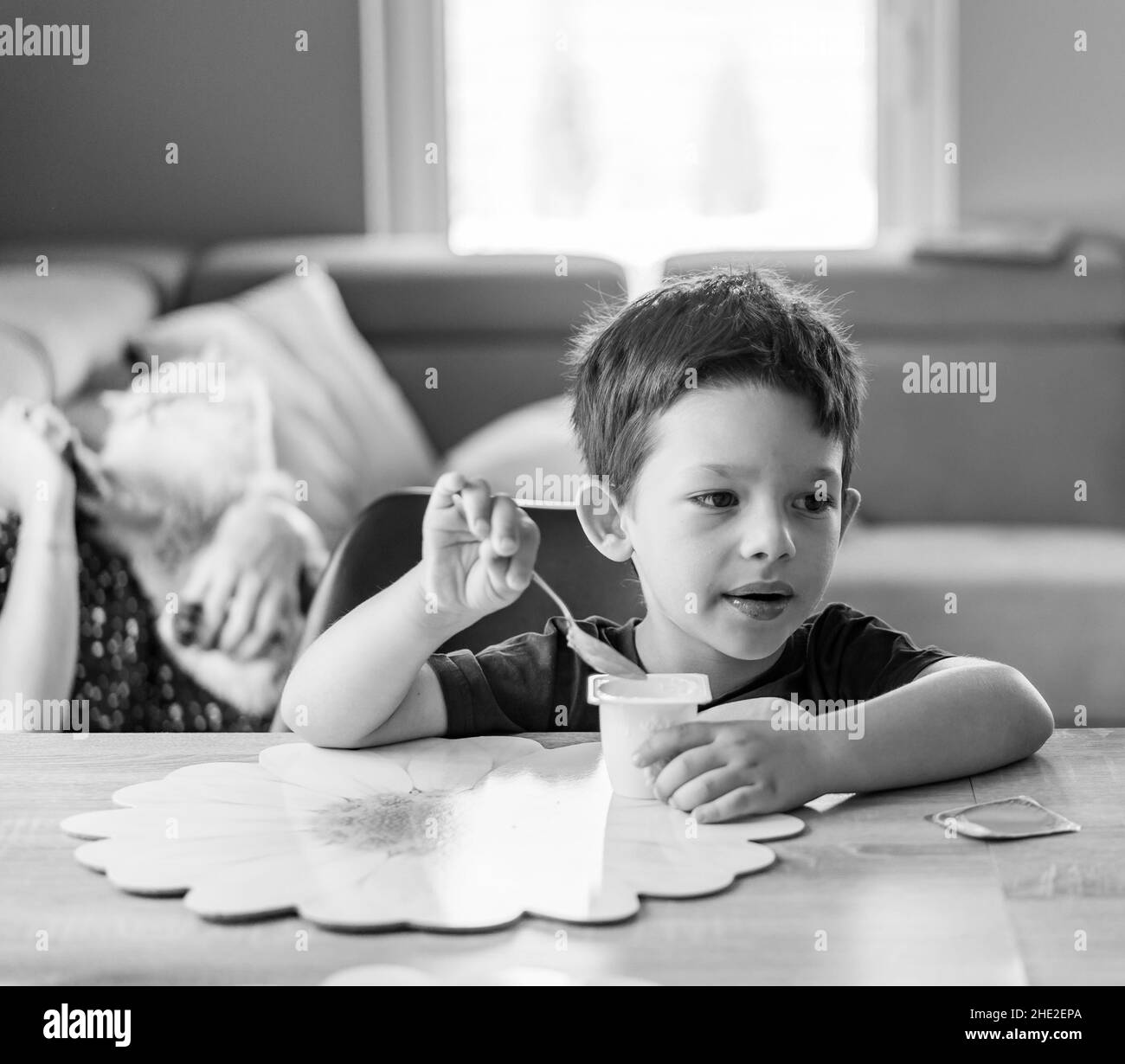 Nahaufnahme eines Jungen, der einen Joghurt isst, während seine Mutter auf dem Sofa auf einem Hintergrund sitzt Stockfoto