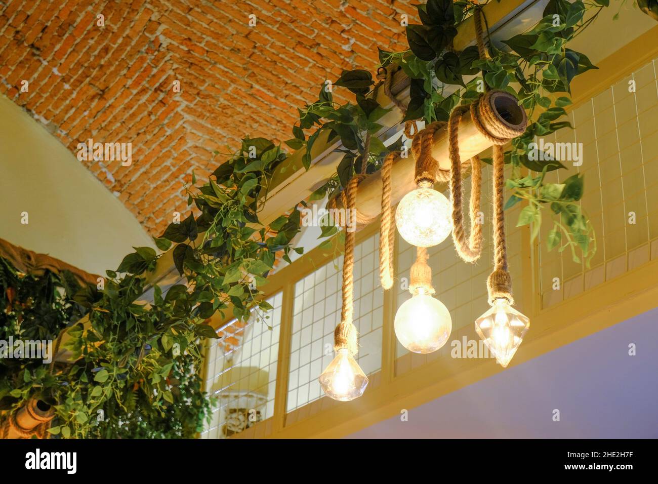 Der dekorative Kronleuchter hängt an Seilen über grünen Pflanzen, Ziegelwände an der Decke. Innenarchitektur. Leichtes Design Stockfoto