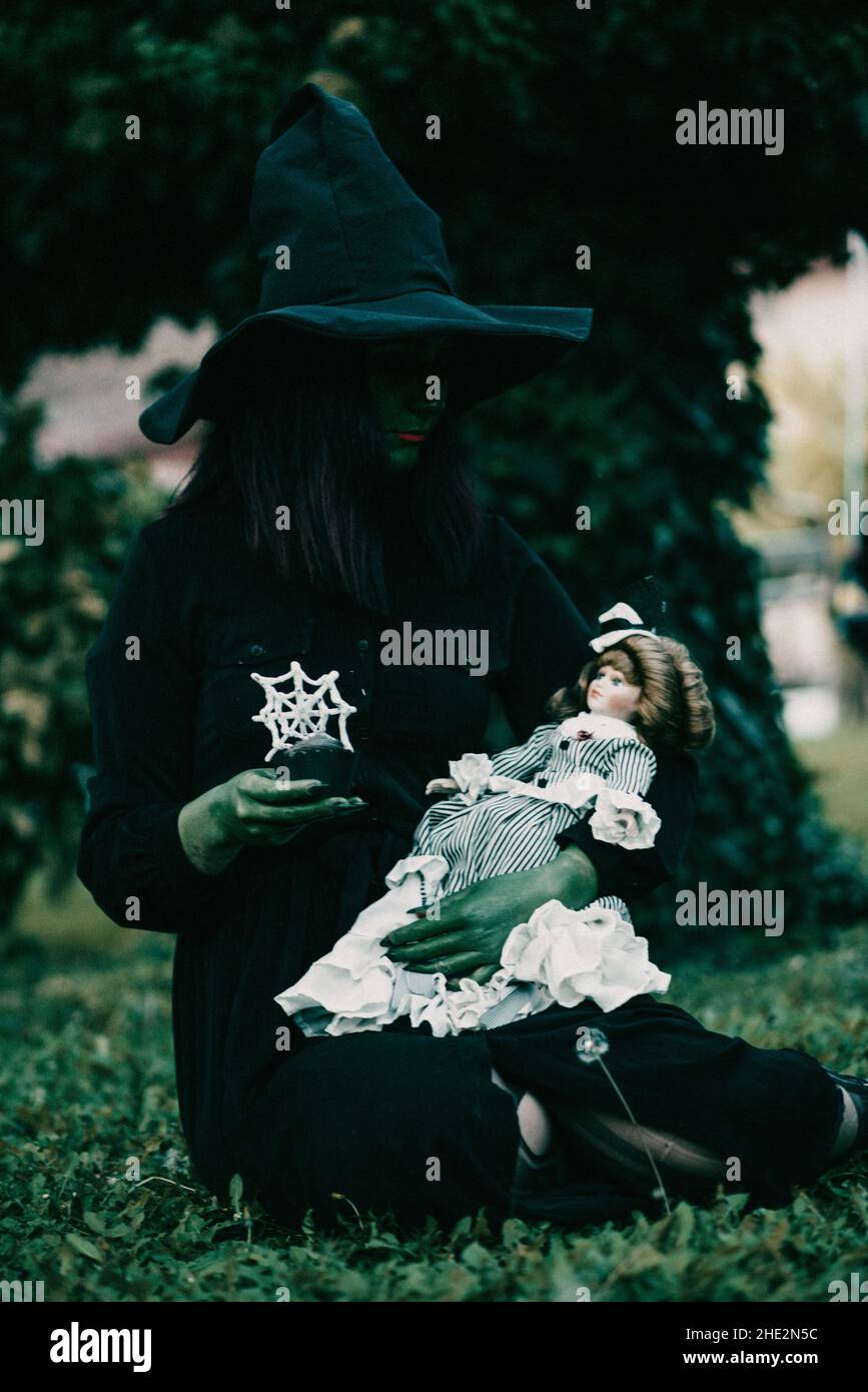Vertikale Aufnahme einer schönen jungen Hexe mit einem Halloween Kostüm und Puppe im Garten Stockfoto